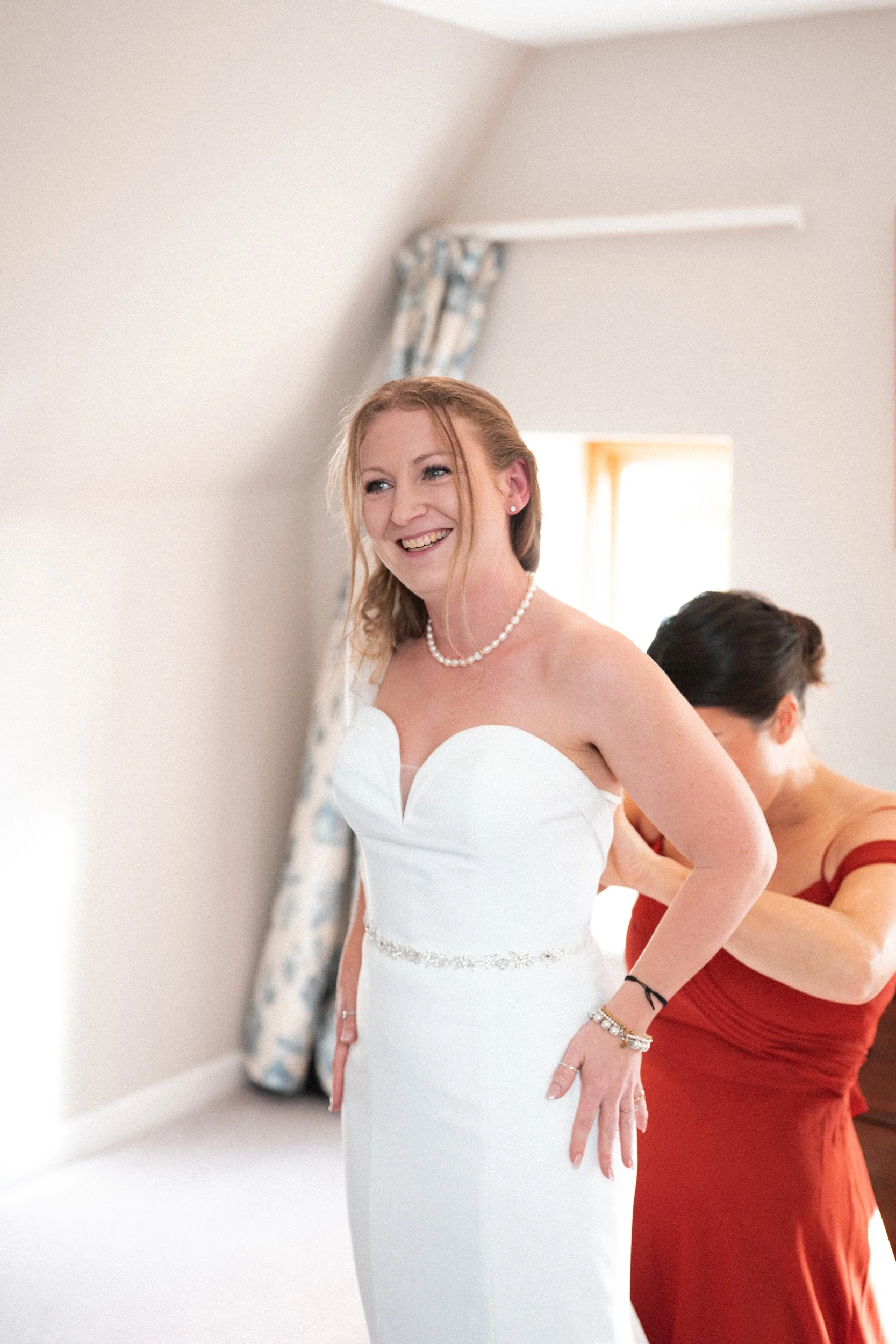 A bride in a white wedding dress smiling while getting ready, with another woman helping her in a room with white walls and a window with floral curtains.