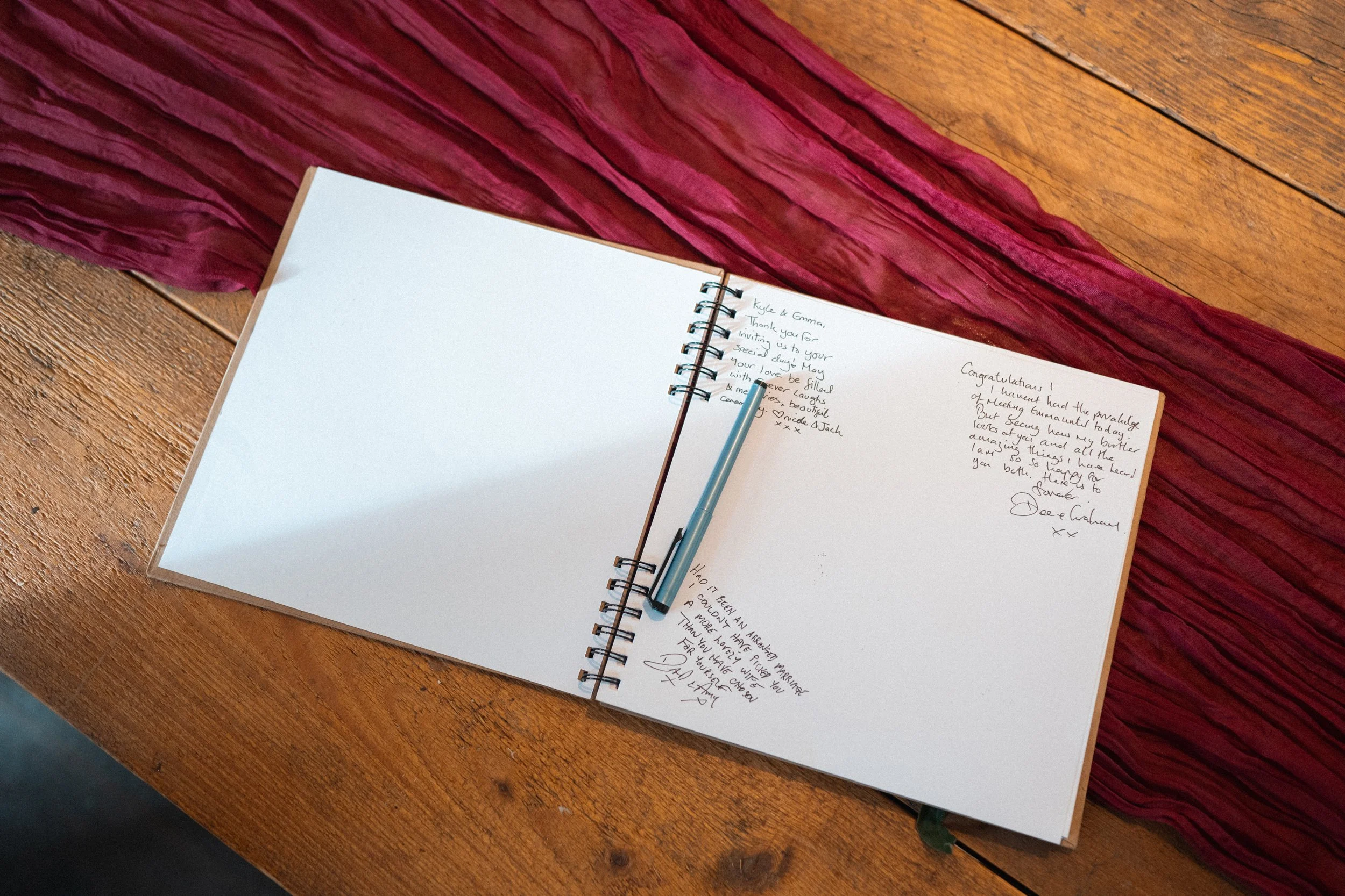 An open guestbook with handwritten messages on a wooden table, with a pen resting on the open pages and a red cloth underneath.