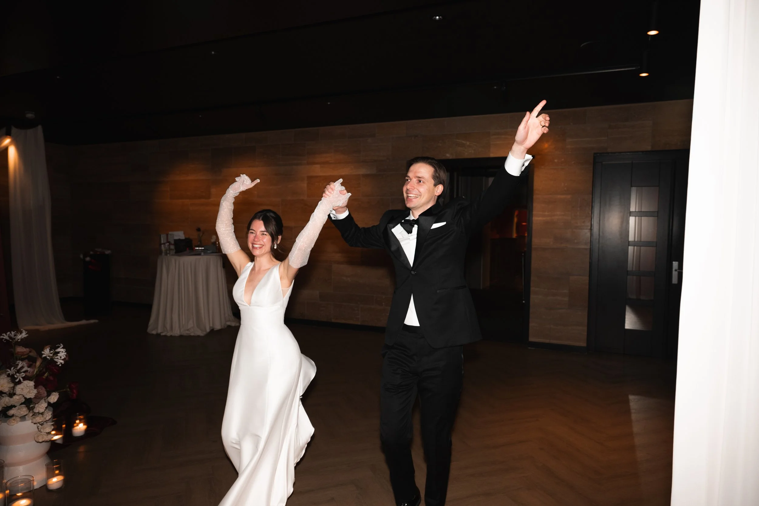 A bride and groom dancing together at their wedding reception, smiling and holding hands with arms raised.