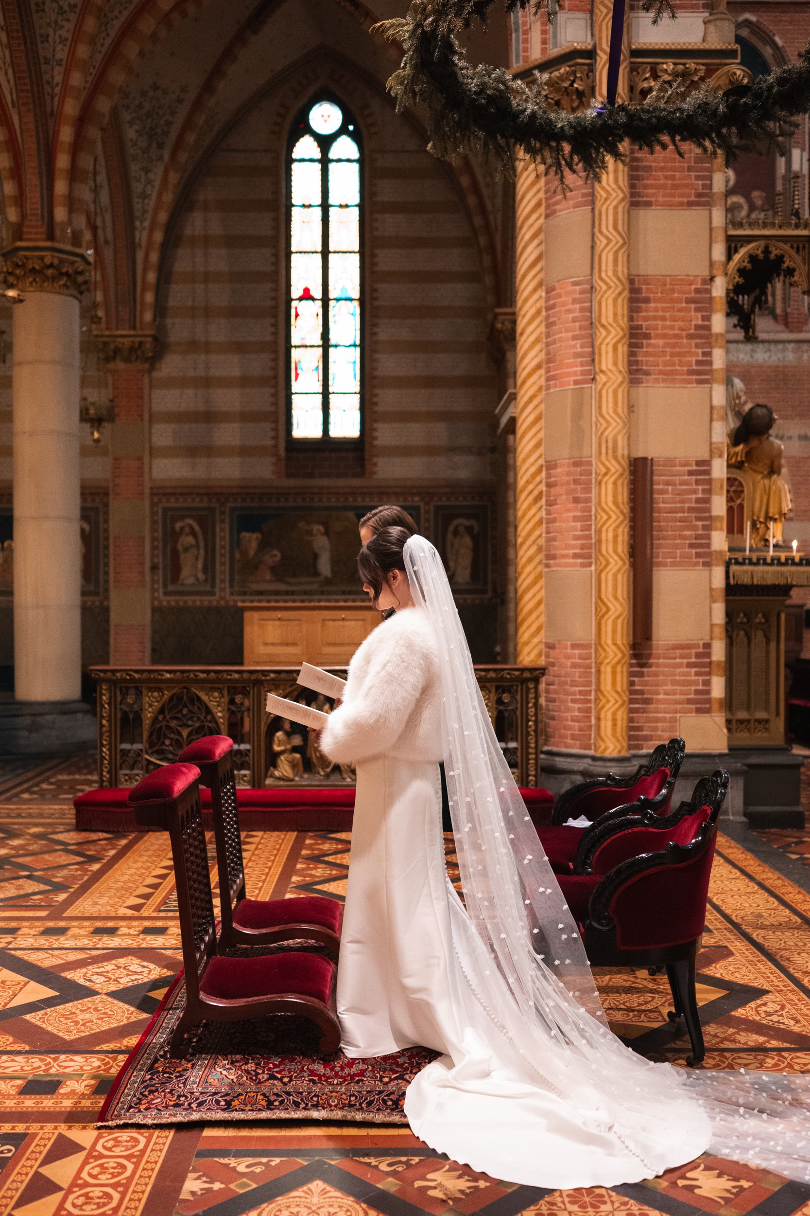 A bride in a white wedding dress and veil standing and reading a book inside a church with stained glass windows and ornate woodwork.