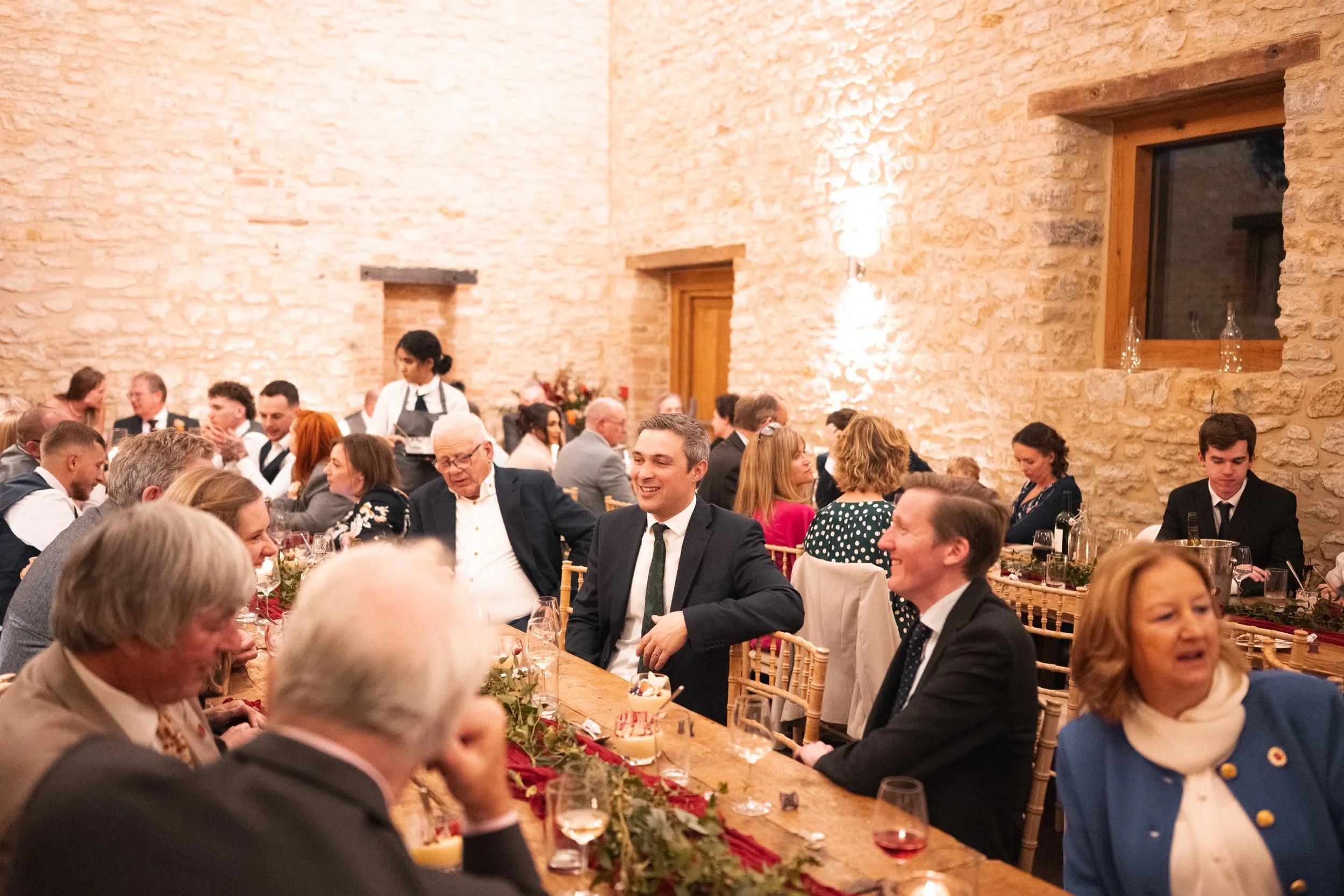 Guests at a formal indoor event sitting at tables with glasses and decorations, engaged in conversations, with brick walls and windows in the background.
