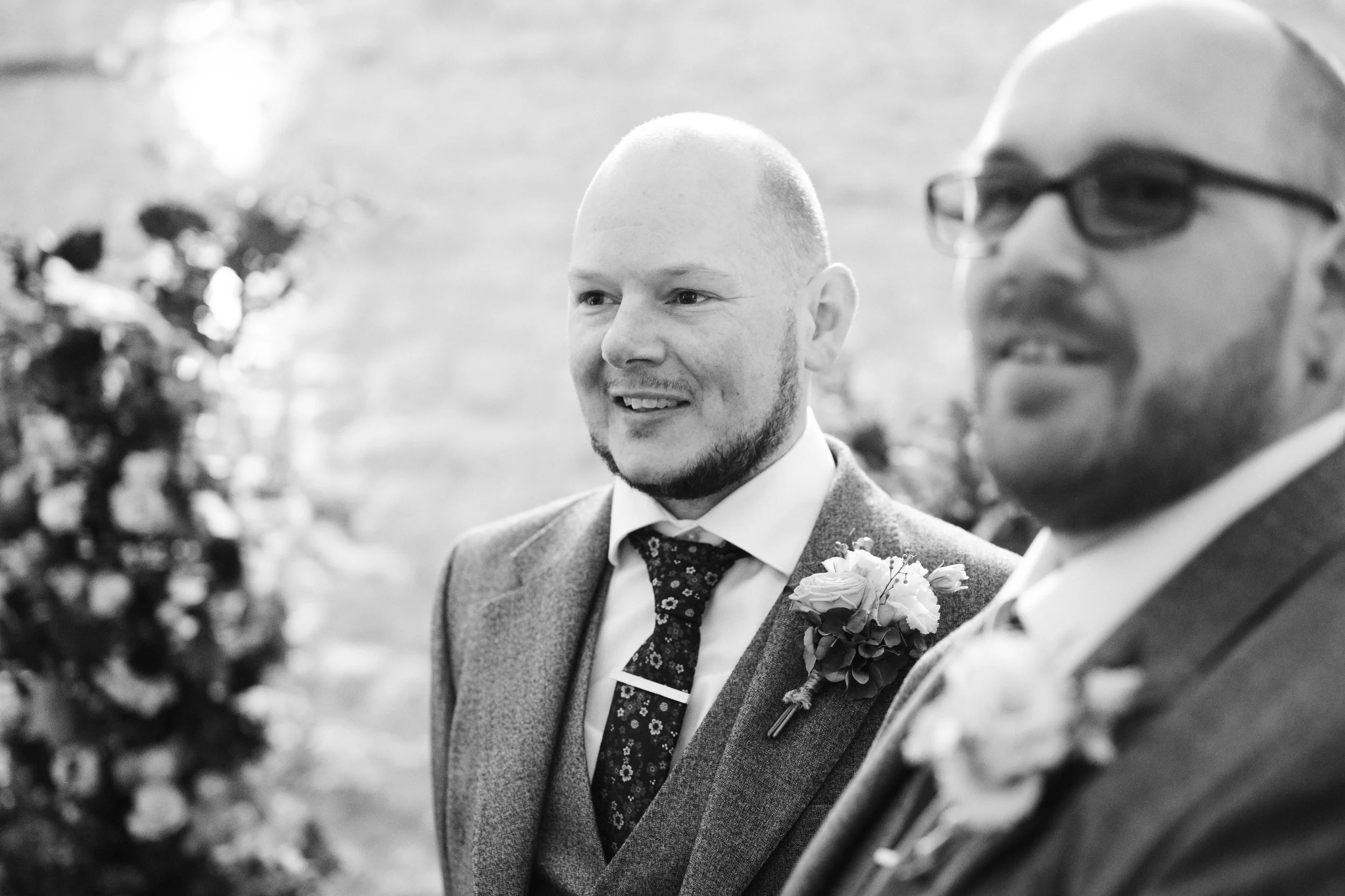 Two men in suits with boutonnieres, at a wedding, smiling. One is bald with a beard, the other has glasses and a beard. The photo is in black and white.