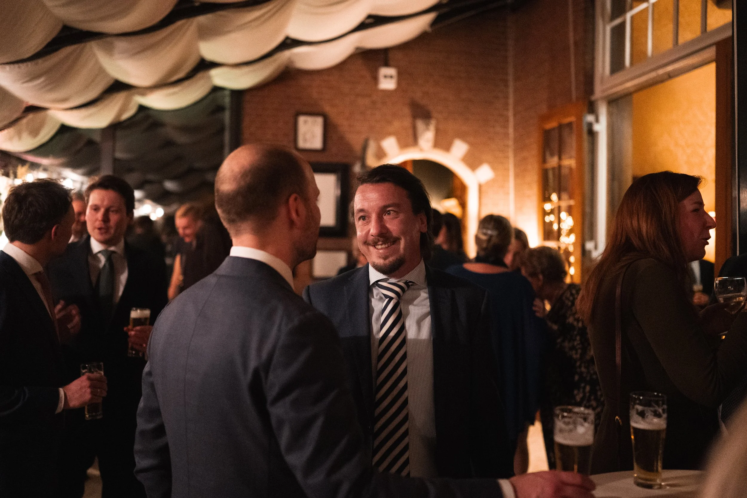 People socializing at a dimly lit gathering, with drinks in hand, indoors with brick walls and decorative ceiling.