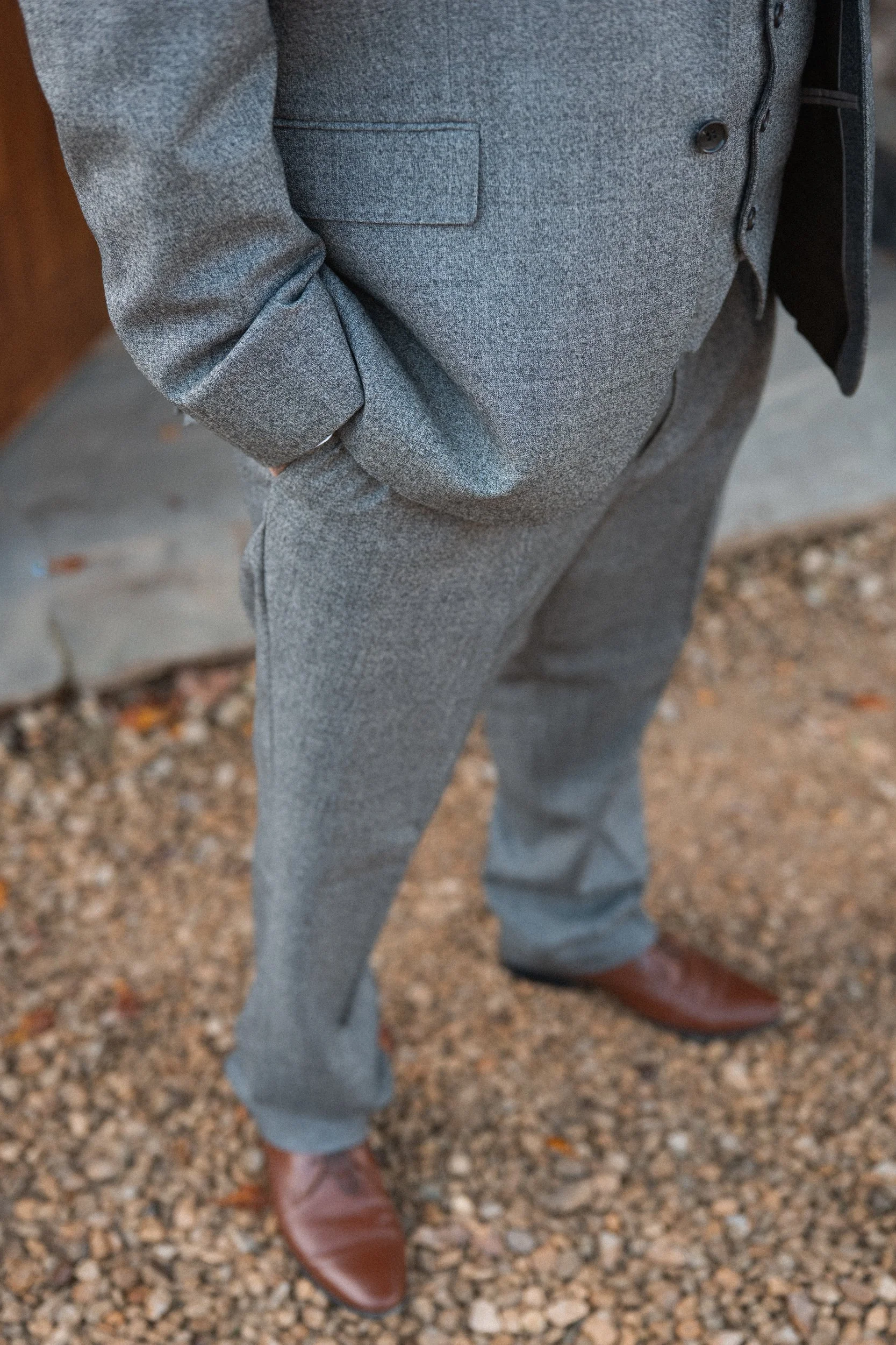 A person wearing a gray suit with their hands in their pockets, standing on gravel.