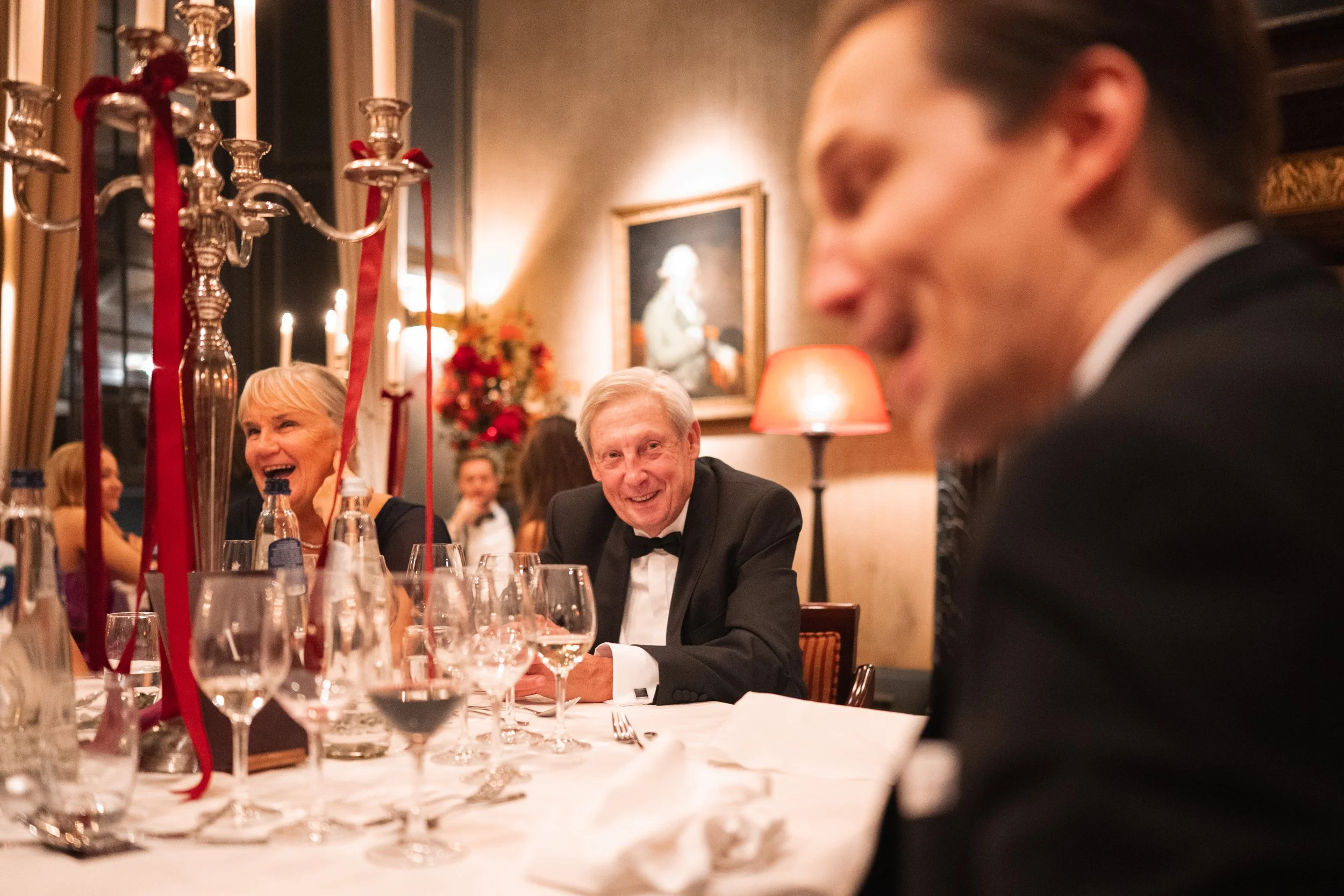 People dressed in formal attire, sitting around a dining table with glasses of wine and water, in a warmly lit restaurant decorated with flowers and paintings, enjoying a celebration.