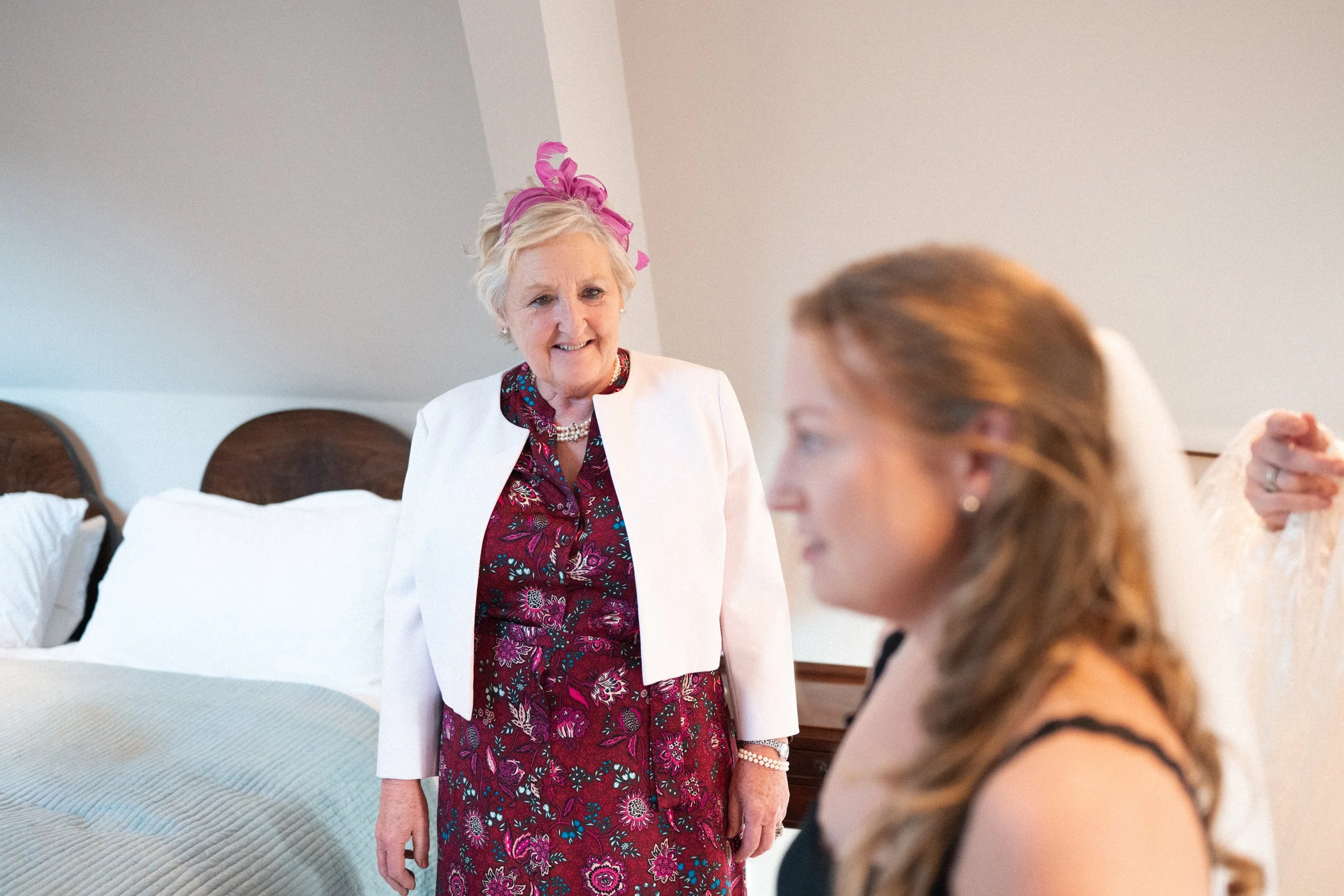An elderly woman in a white blazer and a floral dress stands in a bedroom, looking at a young woman with long hair, dressed in black, who is sitting on a bed with a white headboard.