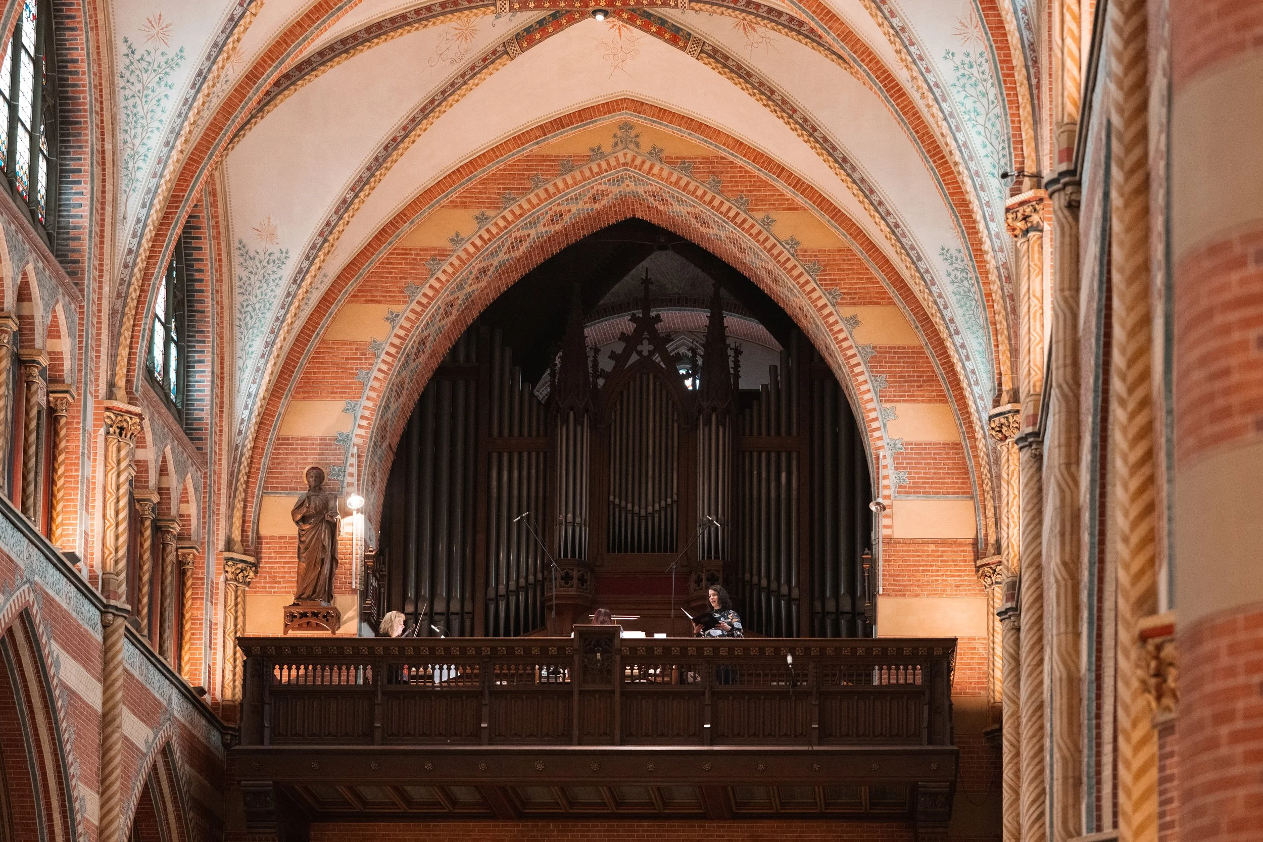 Interior of a church with gothic architecture, featuring high arched ceilings, brick walls, and stained glass windows. There is a statue on the left and a large pipe organ in the background with two people working or performing on a balcony.