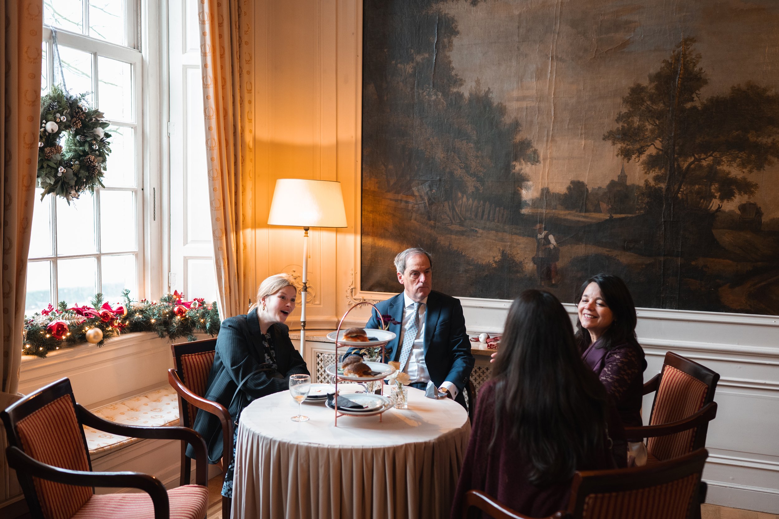 Four people sitting at a round dining table with Christmas decorations, eating and talking in a well-lit house with large windows and a painting on the wall.