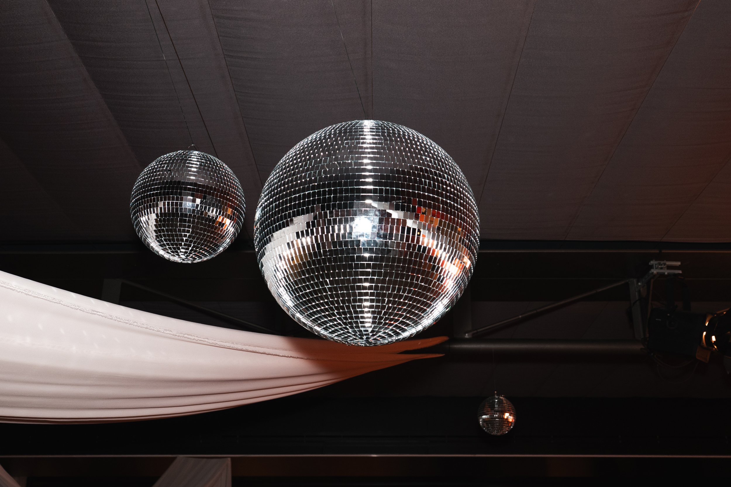 Two large disco balls hanging from a ceiling, reflecting light in a room with dark ceiling panels and white drapery.