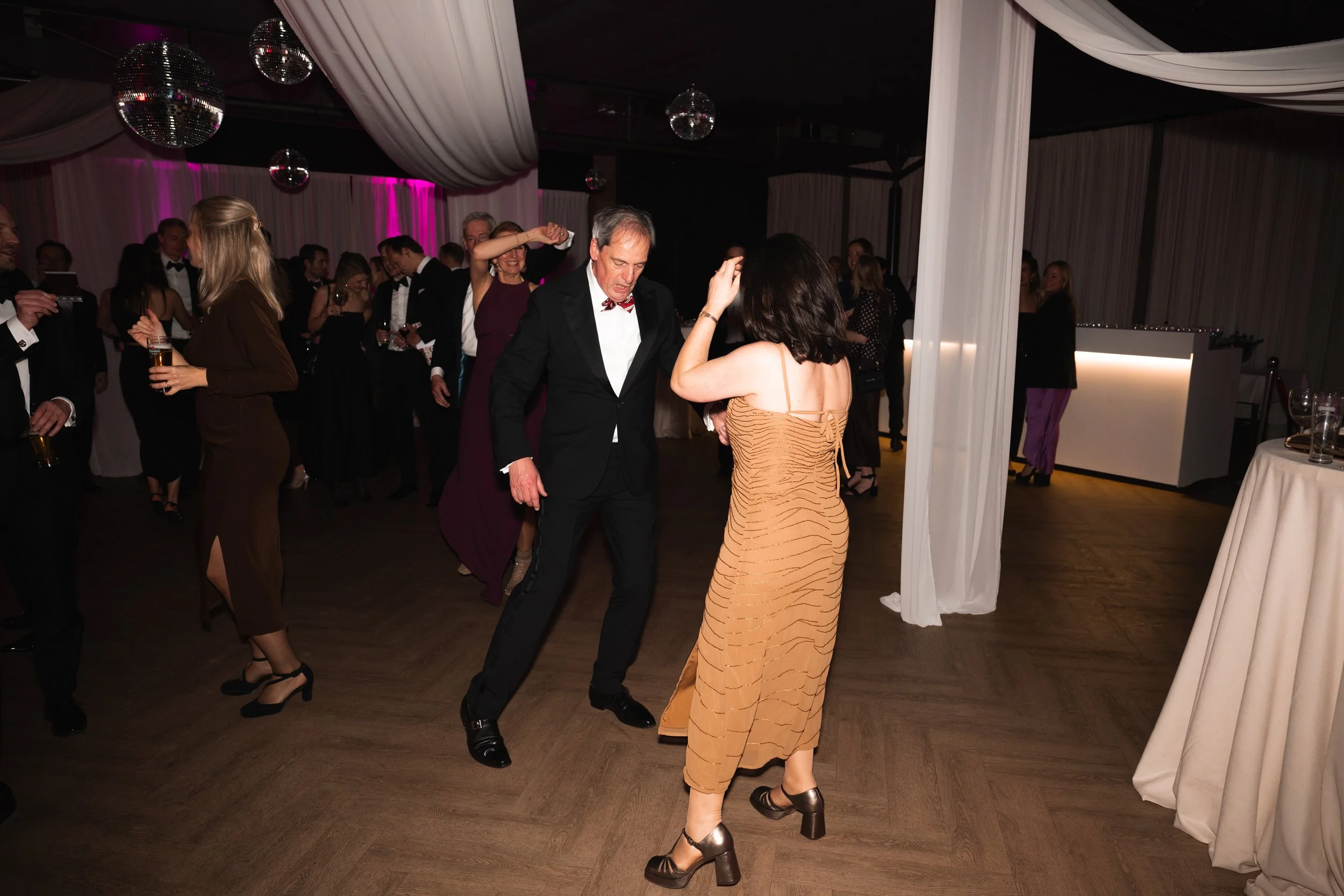 People dancing and socializing at a formal party or wedding reception in a decorated venue with draped fabric and disco balls.