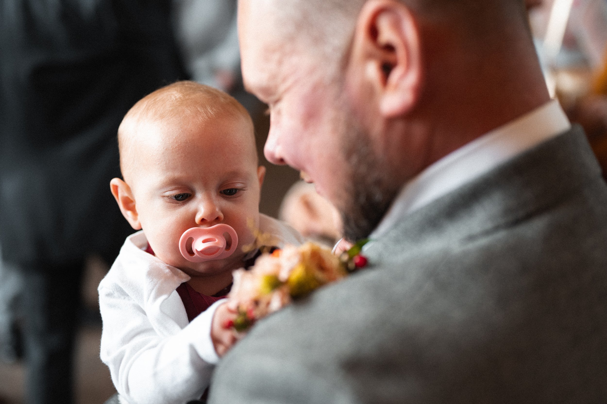 A young child with a pacifier in their mouth is looking at an adult, possibly a father, who is holding them. The man has short hair and a beard, is wearing a suit, and is smiling at the child. The scene appears to be at a social gathering or event.