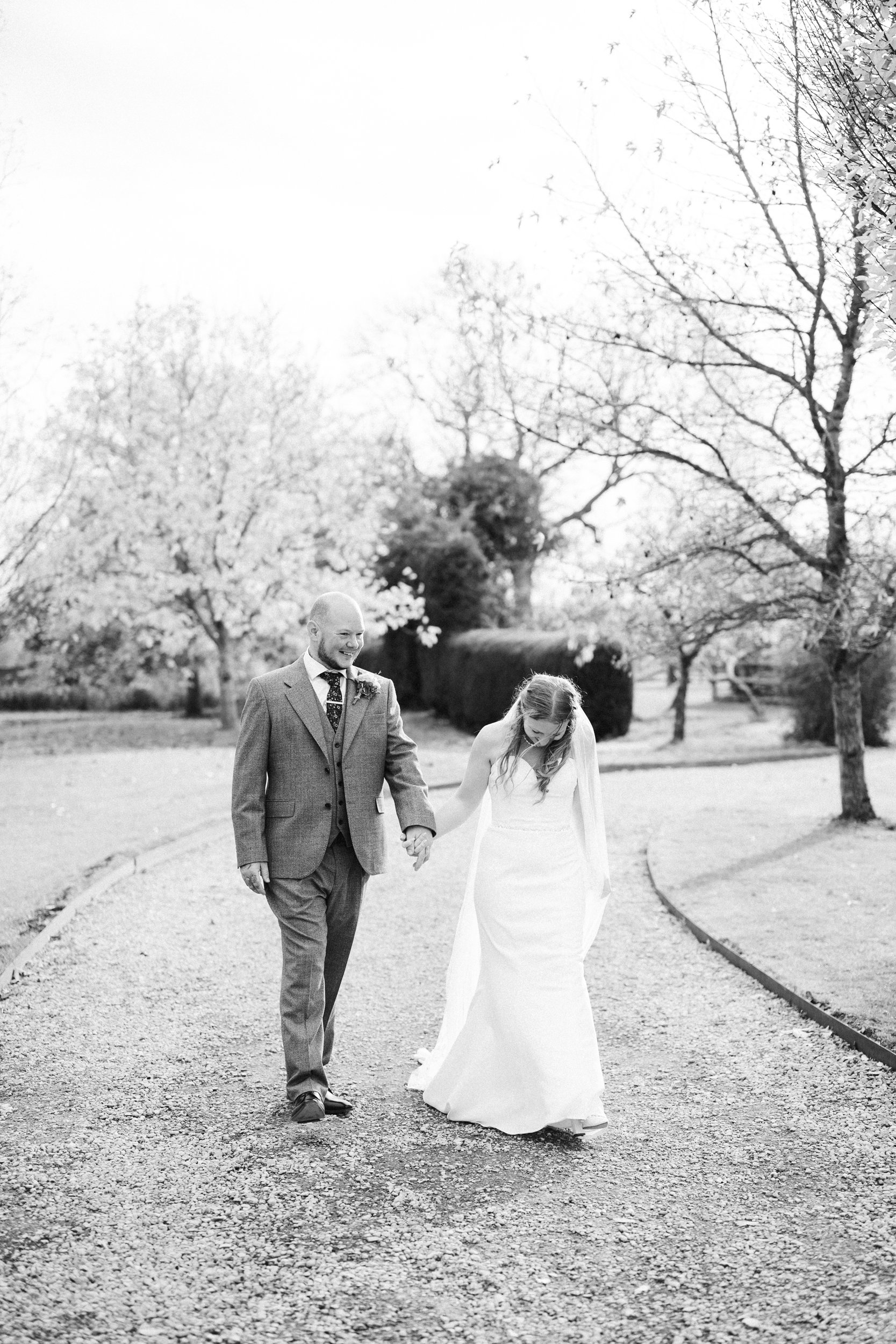 A bride and groom walking hand in hand on a gravel path in a park, smiling and looking at each other, with trees and a clear sky in the background.