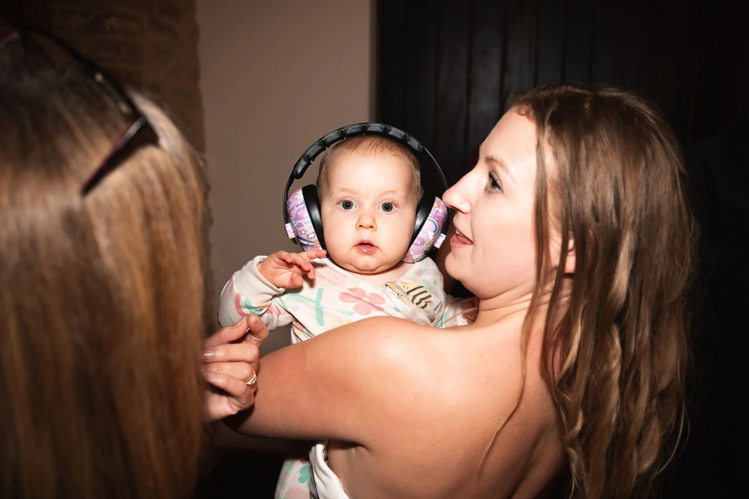 A woman with long, wavy brown hair holding a baby girl with headphones on, looking surprised, with another person partially visible on the left.