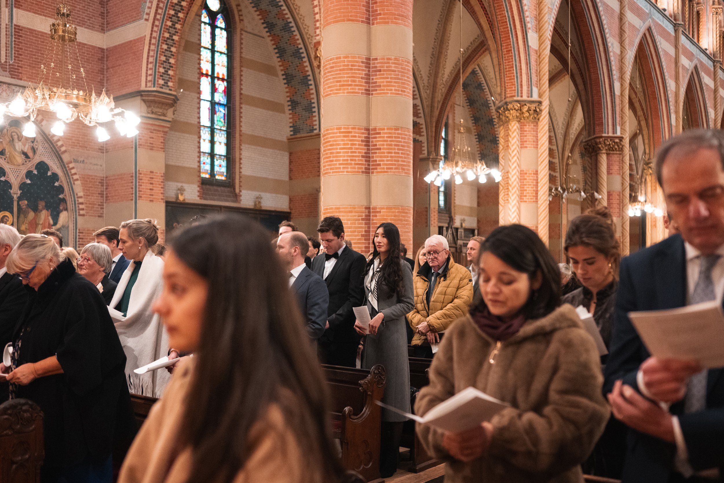 People attending a church service or ceremony inside a church with brick walls, stained glass windows, and chandeliers, standing and holding papers.