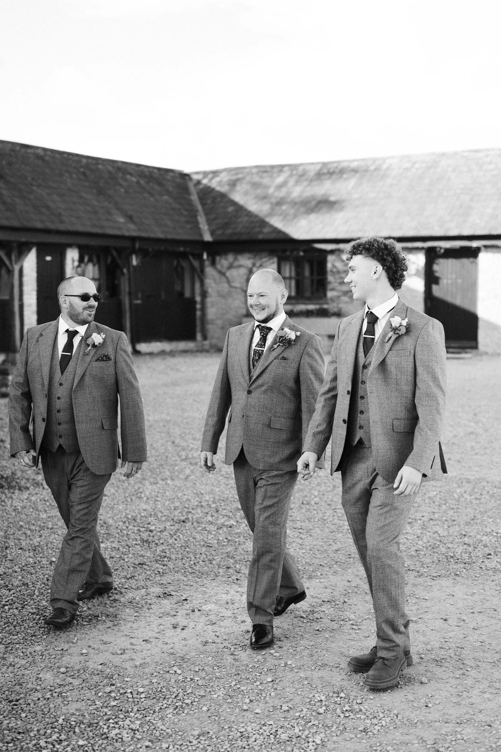 Three men in suits walking outdoors, smiling, in front of a rustic building with a sloped roof, on a gravel surface.