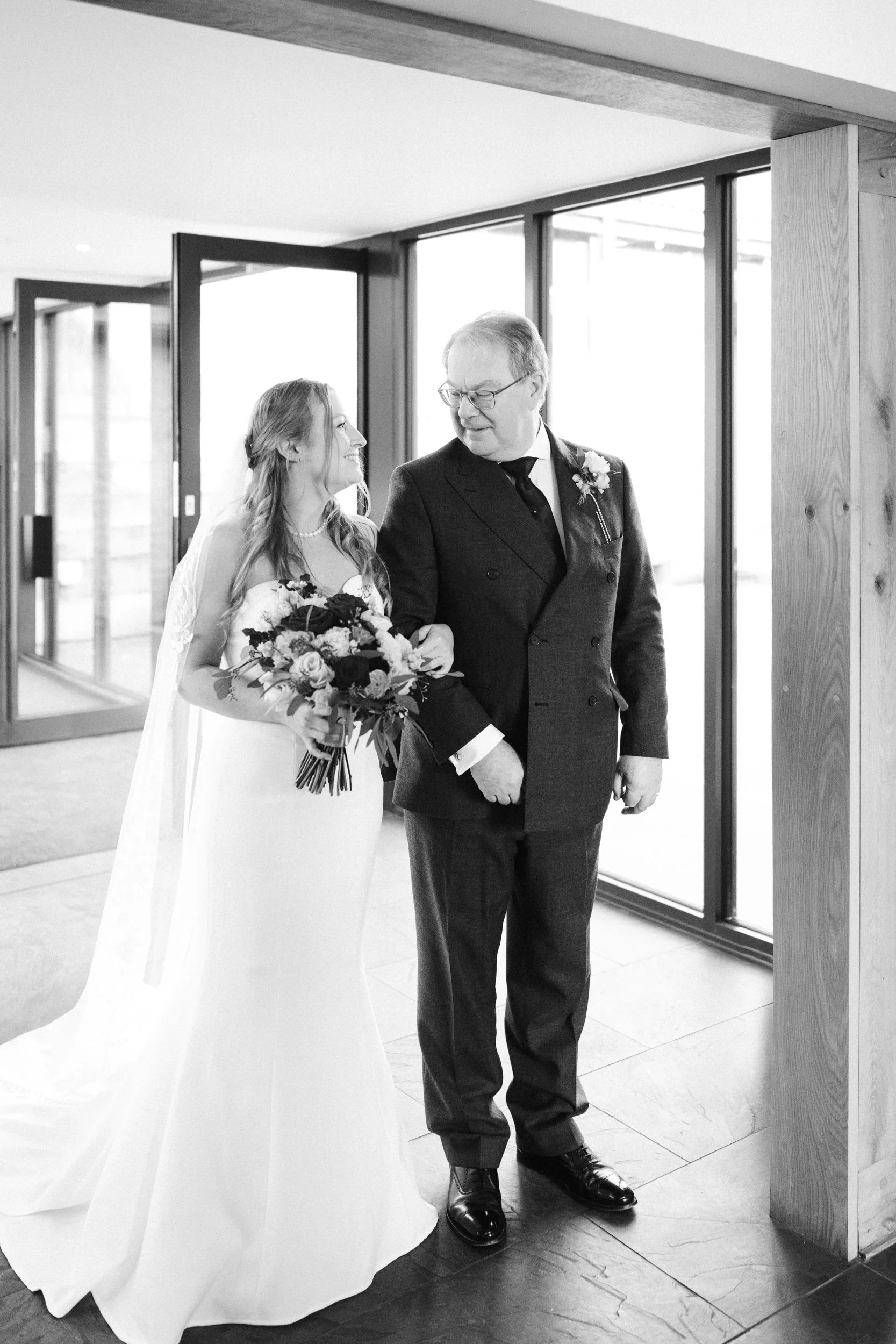 A bride holding a bouquet of flowers, walking arm in arm with an older man, in a bright indoor setting with large windows.