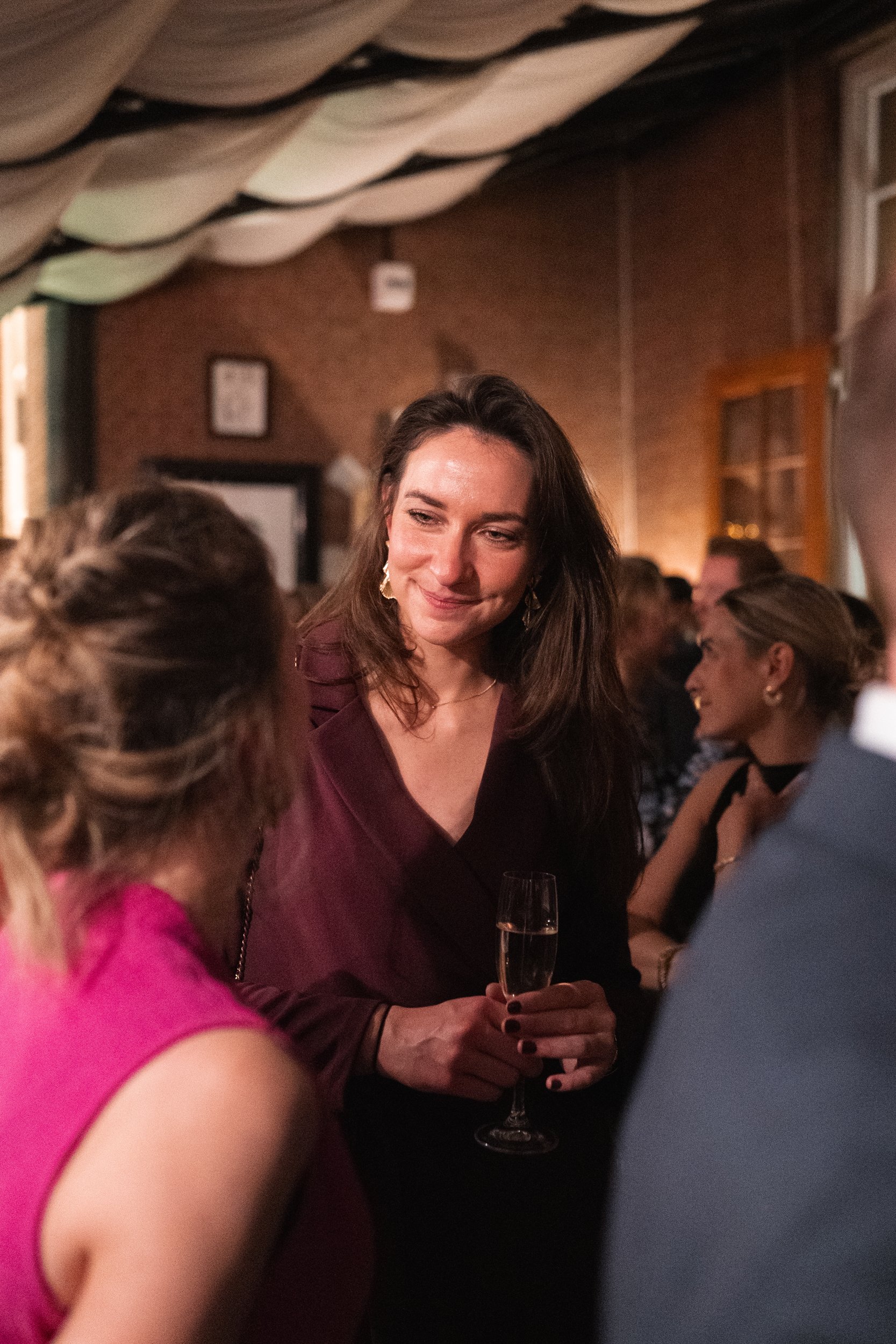 A woman with long brown hair, wearing a dark dress, holding a glass of champagne, smiling and talking to a woman in a pink top at a social gathering in a dimly lit indoor space.