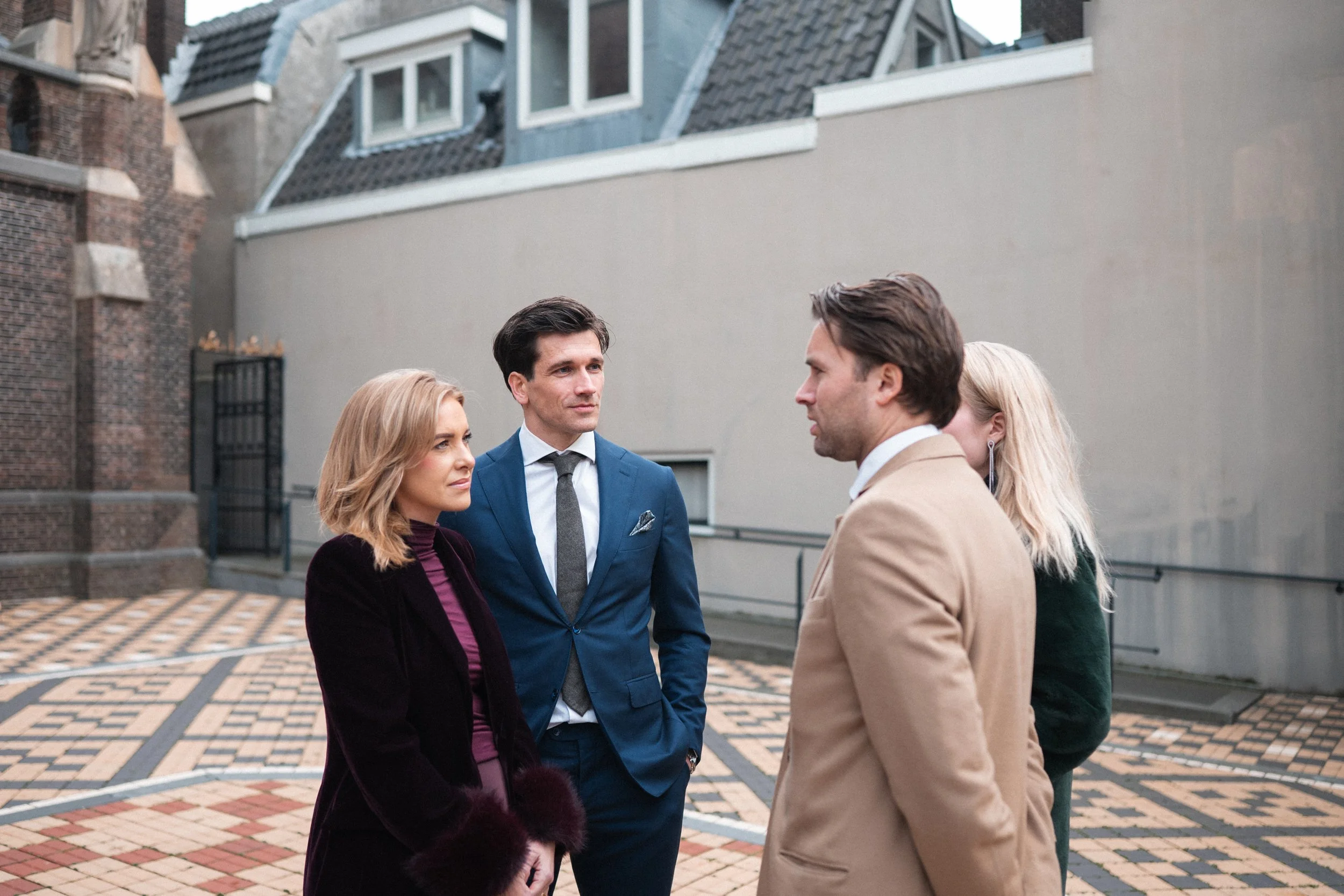 Four people engaged in a conversation outdoors, standing on a patterned brick walkway with buildings in the background. The group includes two men and two women, dressed in formal and semi-formal attire.