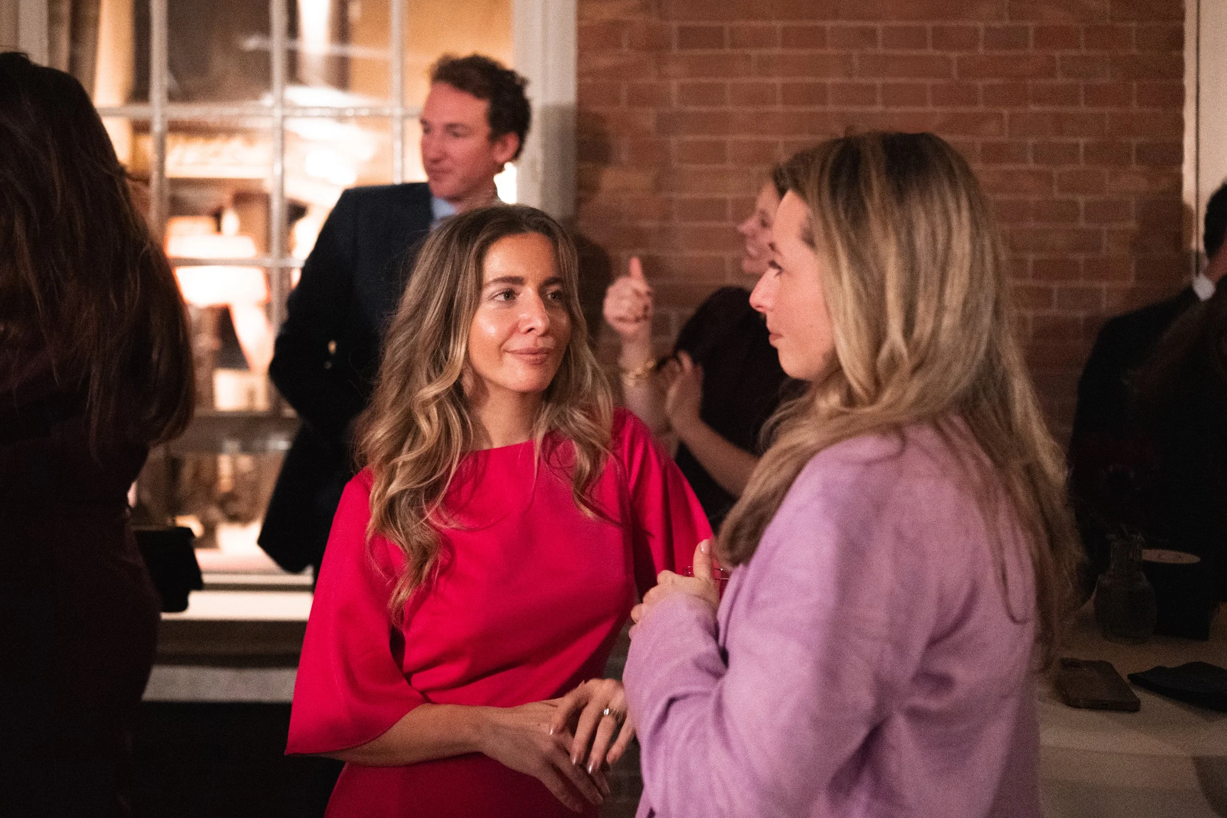 Two women are talking to each other at a social event in a warmly lit room with brick walls. One woman is wearing a bright pink dress, and the other is wearing a light purple blazer.