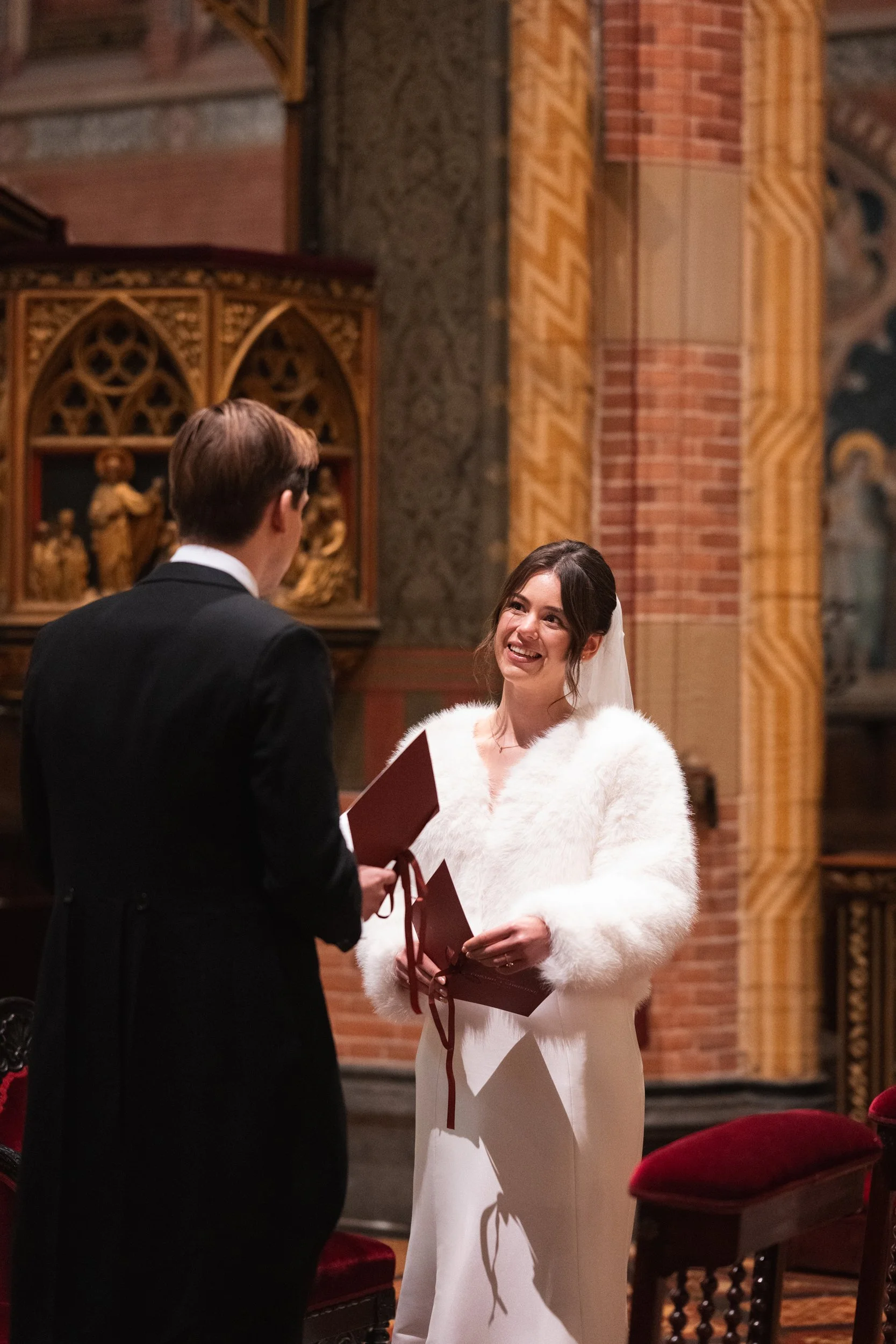 A woman in a white dress and faux fur coat exchanges vows with a man in a black tuxedo inside a church with ornate brick and gold decorations.