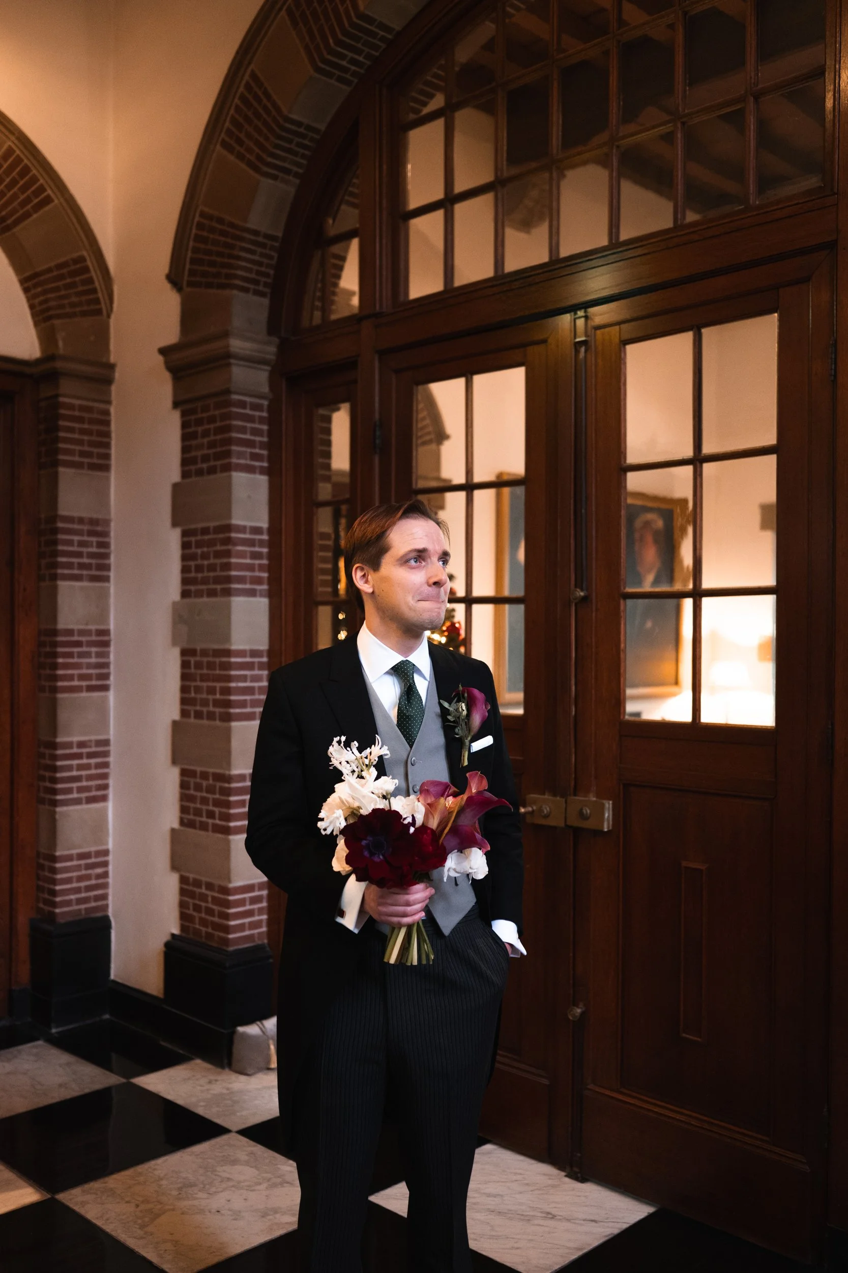 A man dressed in formal attire holding a bouquet of flowers, standing indoors near wooden doors with large glass panes, with a brick wall and paintings visible in the background.