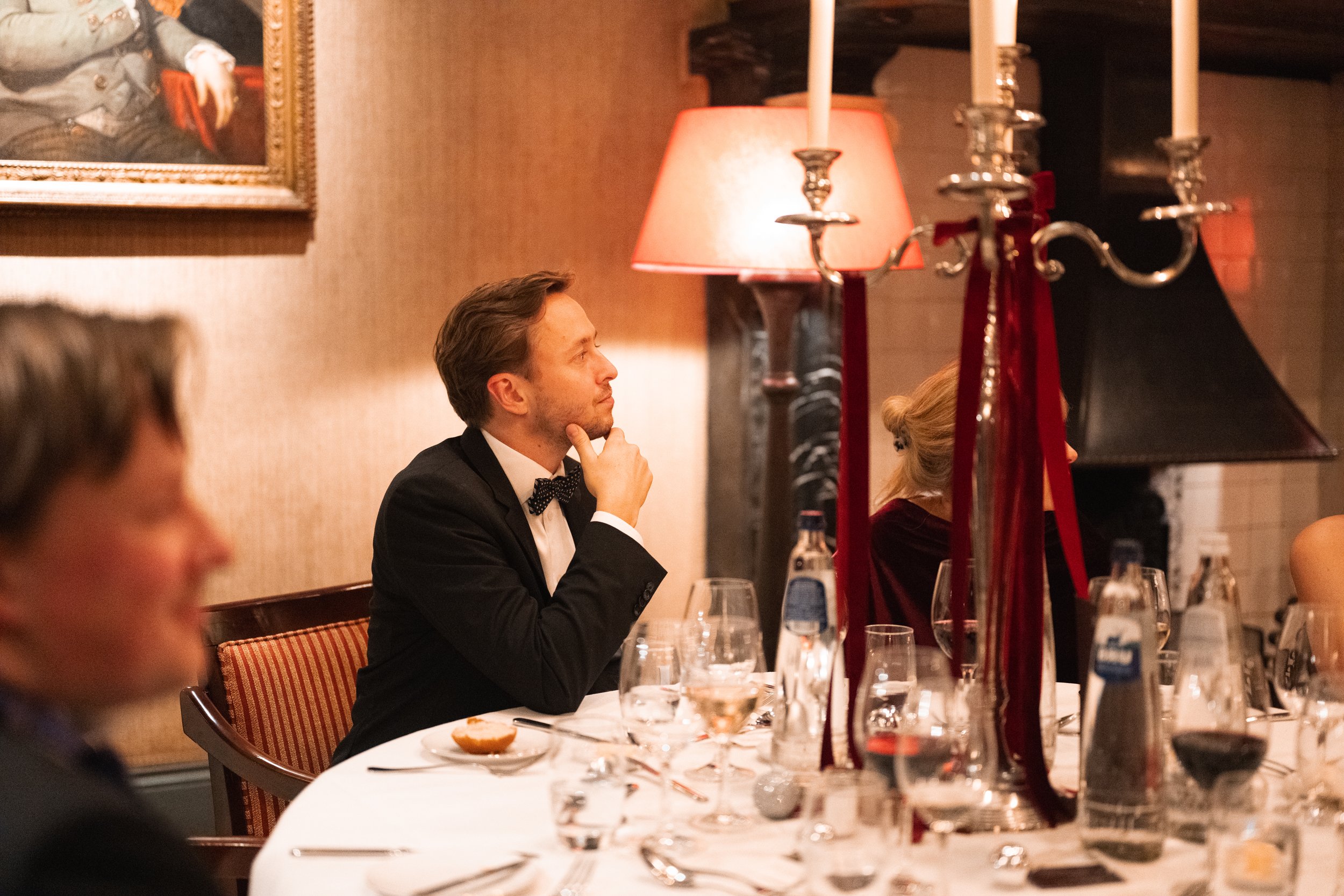Man in a tuxedo sitting at a dinner table, resting his chin on his hand, in an elegant dining room.