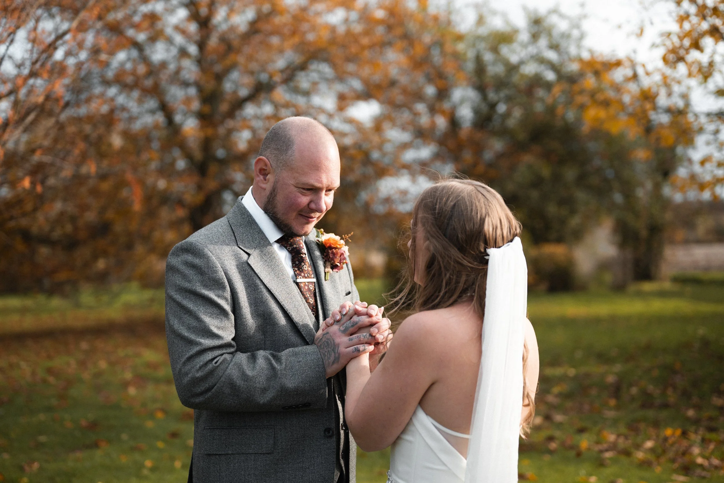 A groom and bride holding hands during their outdoor wedding ceremony in autumn, with orange and yellow trees in background.