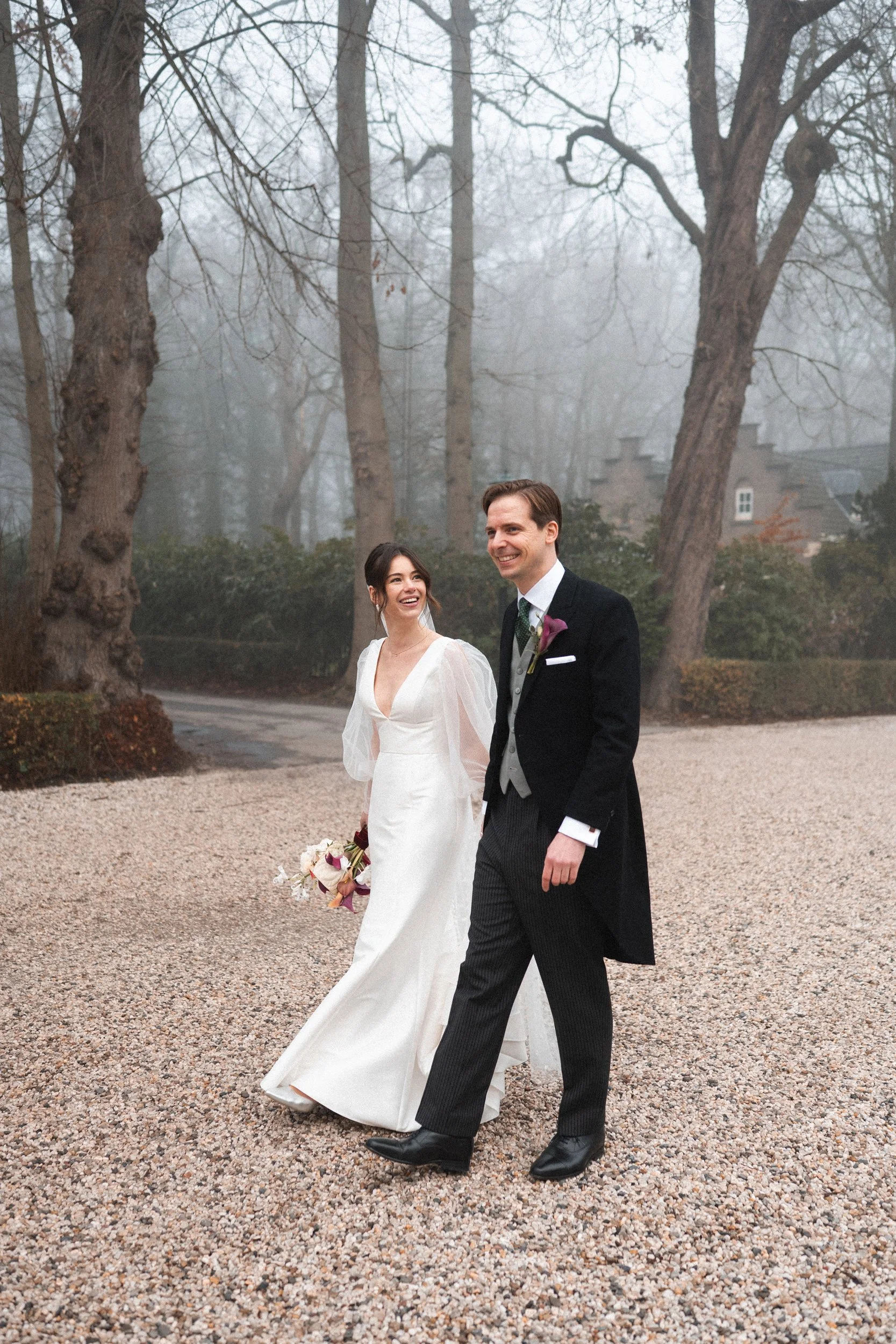 A bride in a white wedding gown and a groom in a black tuxedo walk together outdoors on a gravel path, smiling, with leafless trees and a foggy background.