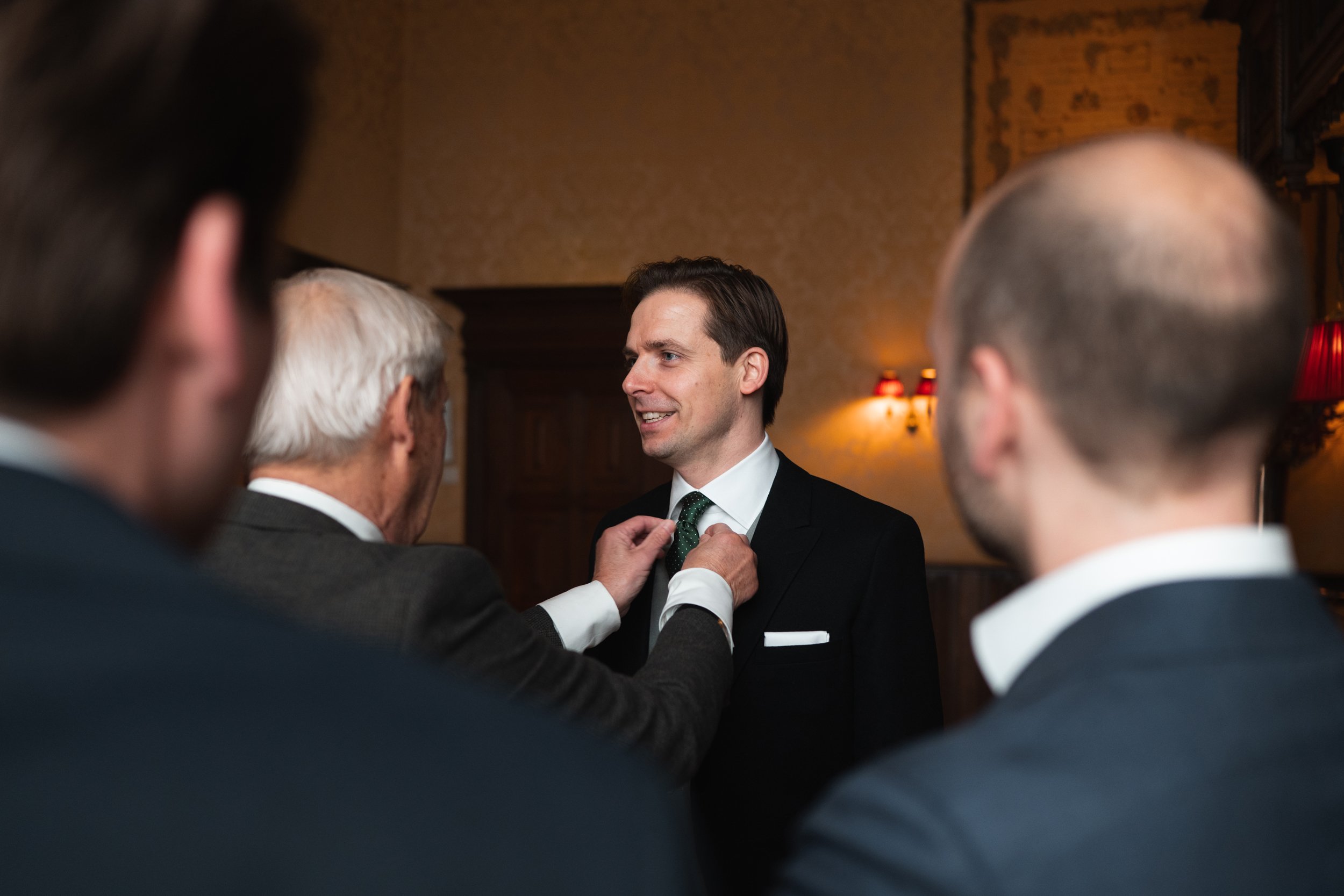 A man in a black suit is being helped with his green tie by an older man while two other men watch, all dressed in suits, in an elegant room with warm lighting.