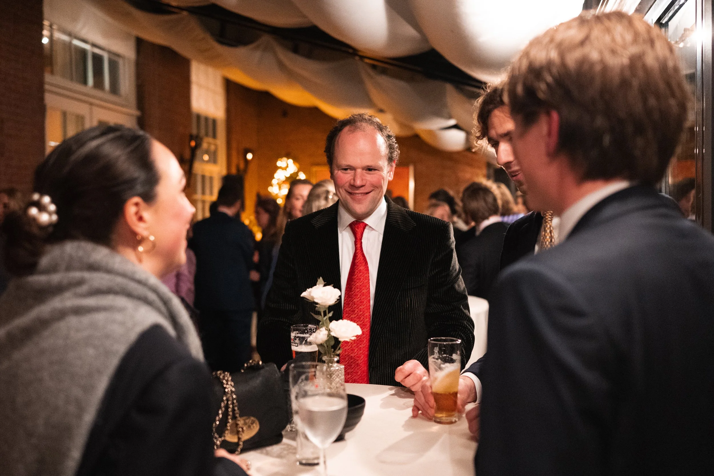 A group of four adults having a conversation at a social event, standing around a table with drinks, in a warmly lit indoor setting decorated with flowers and string lights.