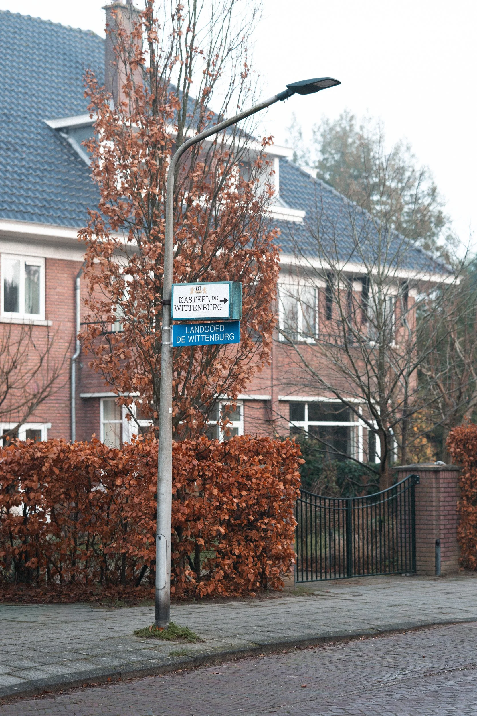 Street scene with a street sign that reads "KAISERLEIN DE WITTENBURG" with an arrow and another sign below it that says "LANDGOED DE WITTENBURG". There is a house with blue roofing tiles, trees with autumn leaves, a black gate, and a brick sidewalk.