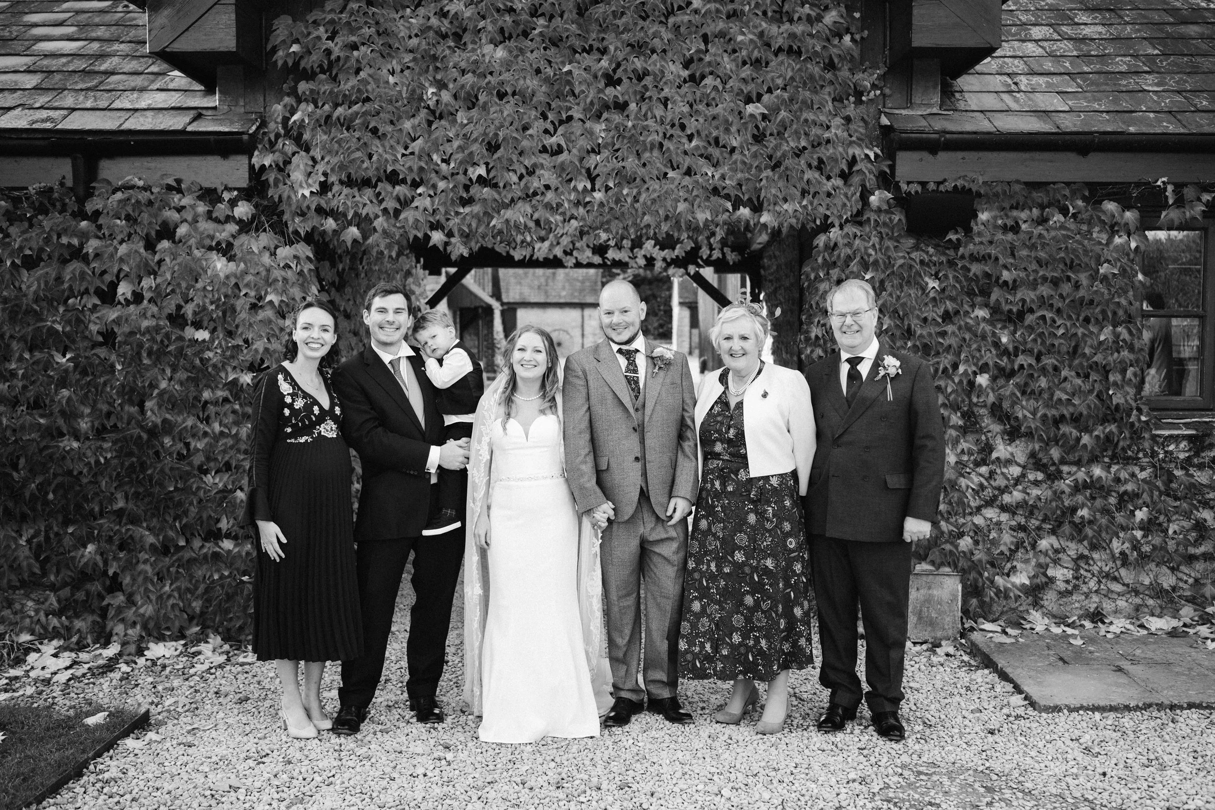 Group of people at a wedding, standing outdoors in front of a leafy wall, dressed in formal attire, smiling for the camera.
