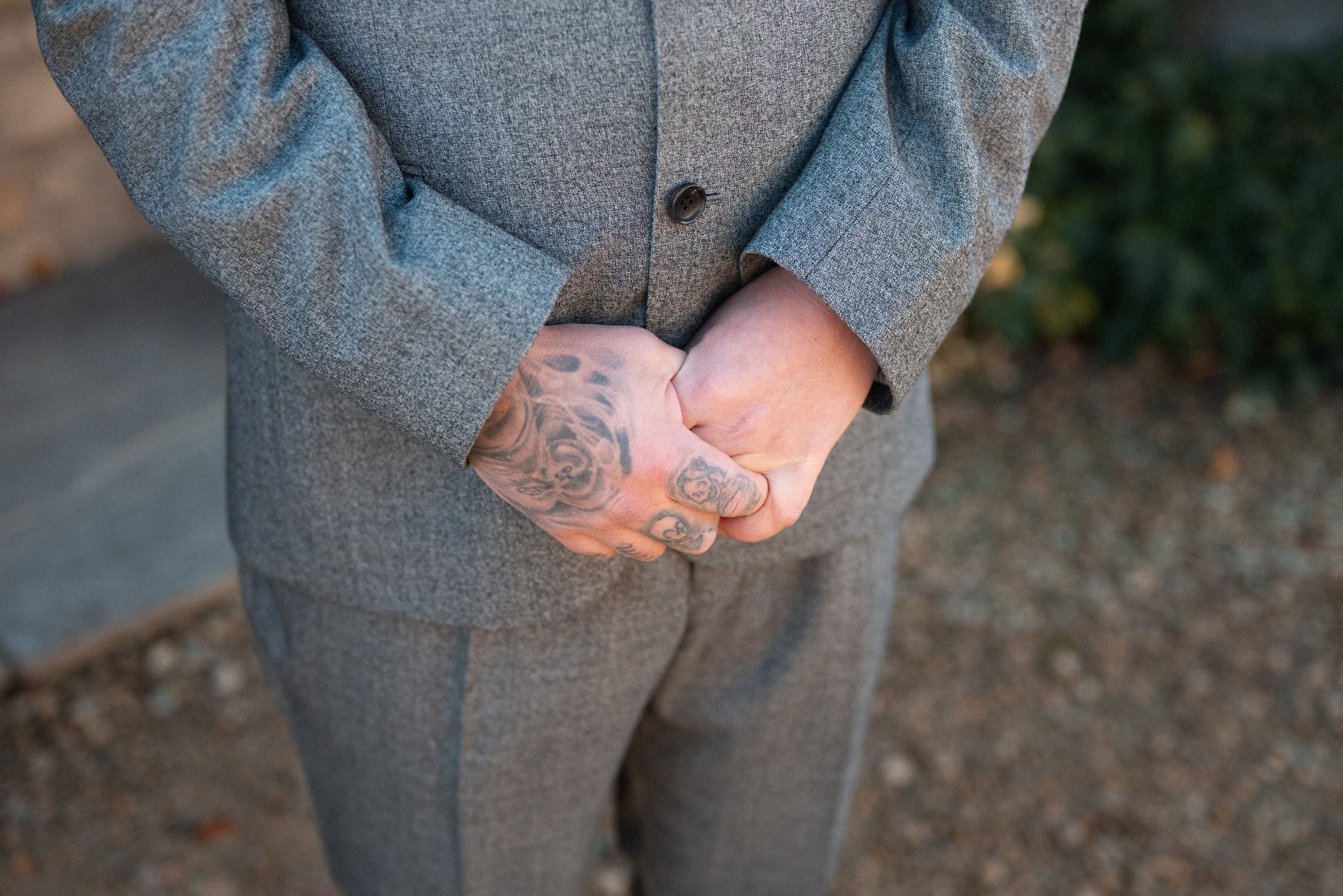 Person wearing a gray suit with hands clasped in front, showing tattoos on the hands and forearms, outdoors with blurred greenery and ground in the background.