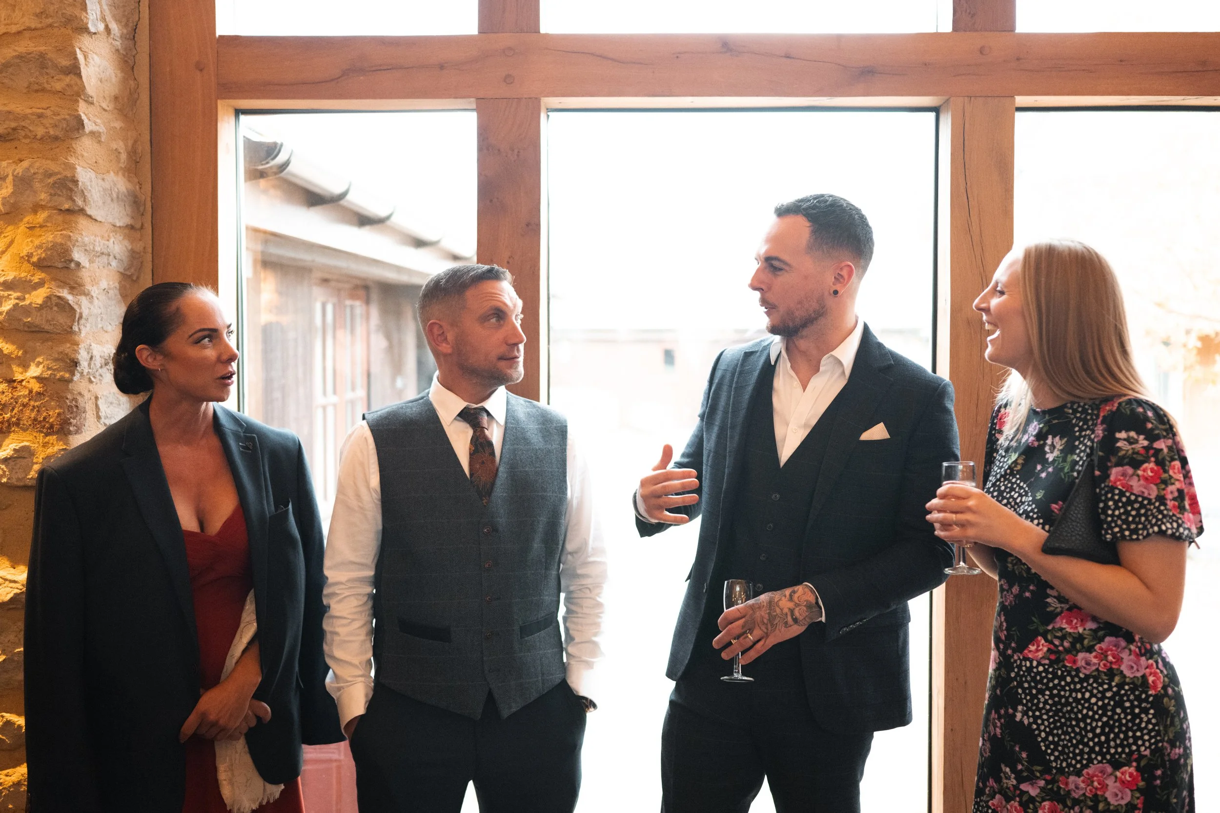 Four people in formal attire conversing at a social event near a large window, with wine glasses in hand.
