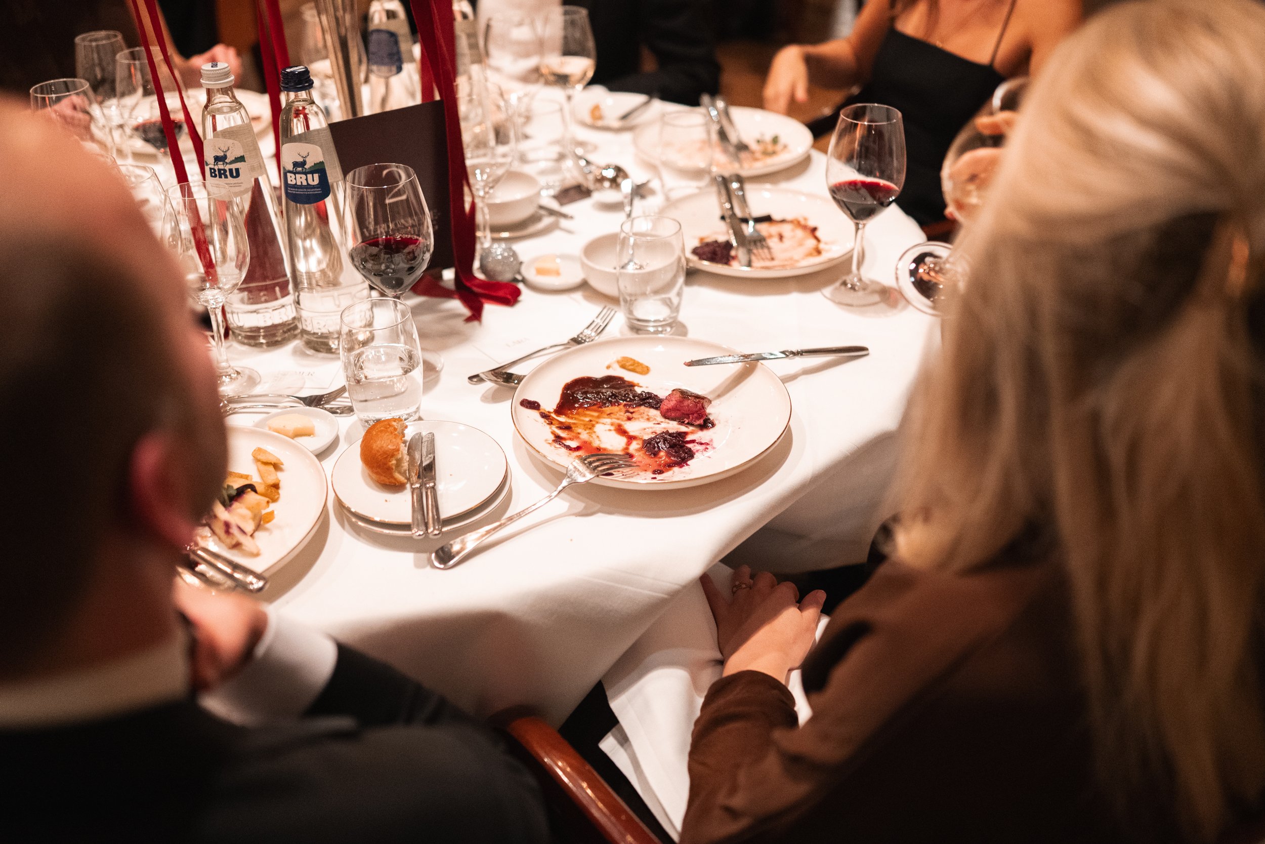 A dinner table with empty plates, wine glasses, water bottles, and remnants of food, indicating a meal has finished, with some guests still seated around the table.