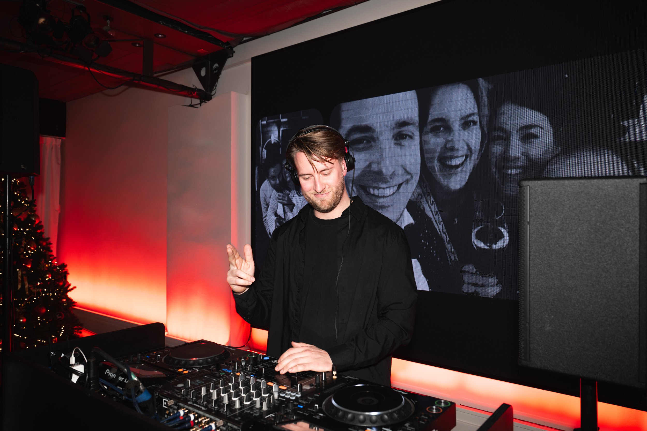 A DJ standing at a mixing console wearing headphones and making a peace sign with his fingers, with a large screen behind him displaying a black-and-white photo of smiling people and a Christmas tree with lights to the side.