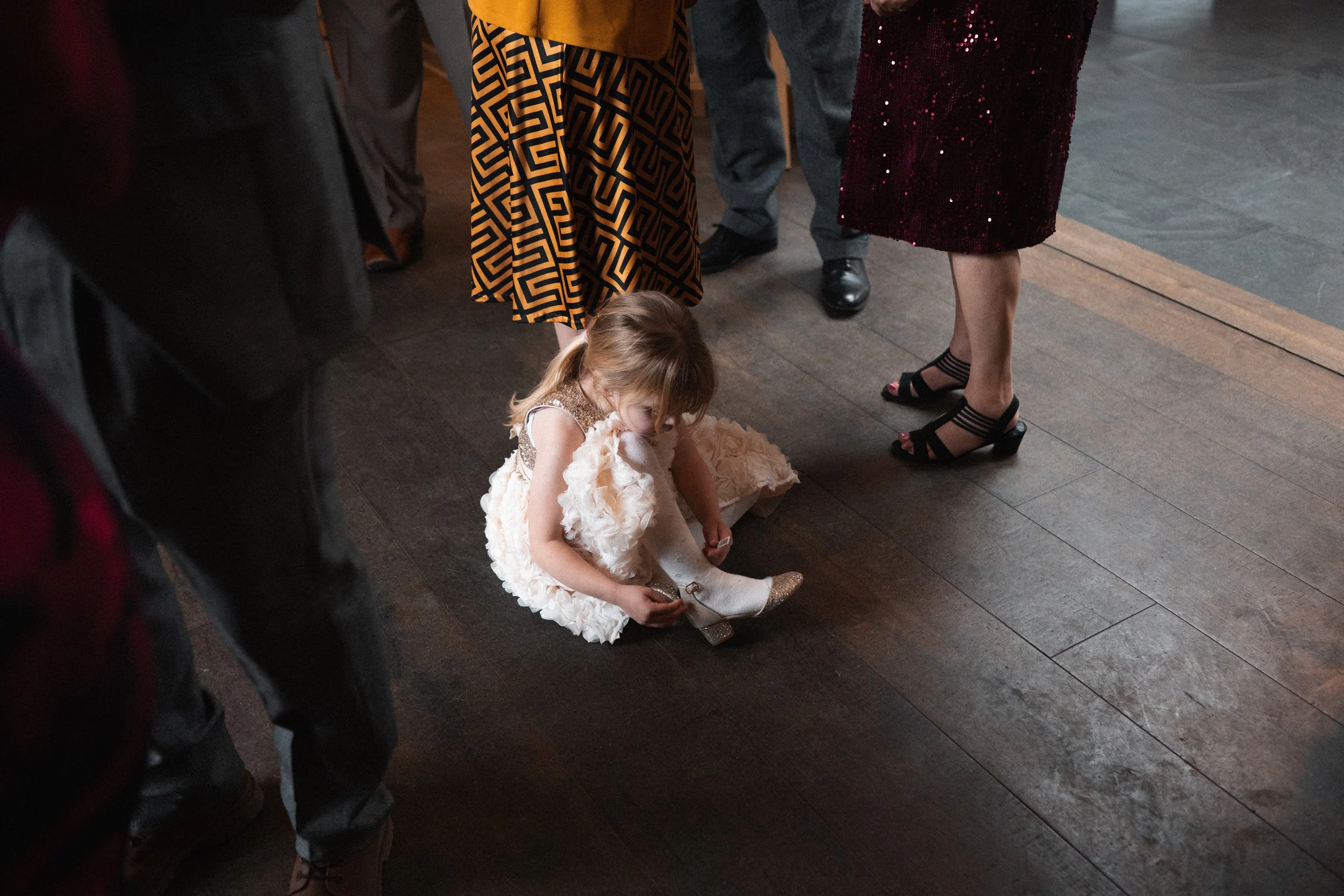 A young girl in a fancy white dress with knee-high boots is sitting on a dark wooden floor, tying the boots. She is surrounded by adults dressed in formal attire, including women in high heels and a woman in a sequined dress.