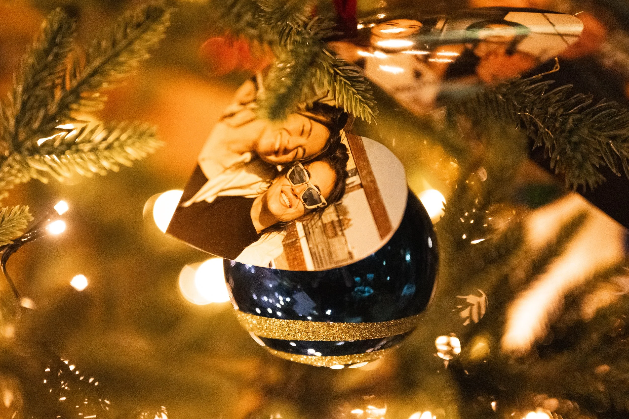 Decorated Christmas tree with a reflective ornament showing a photo of two women smiling and wearing sunglasses, surrounded by blurred warm lights.
