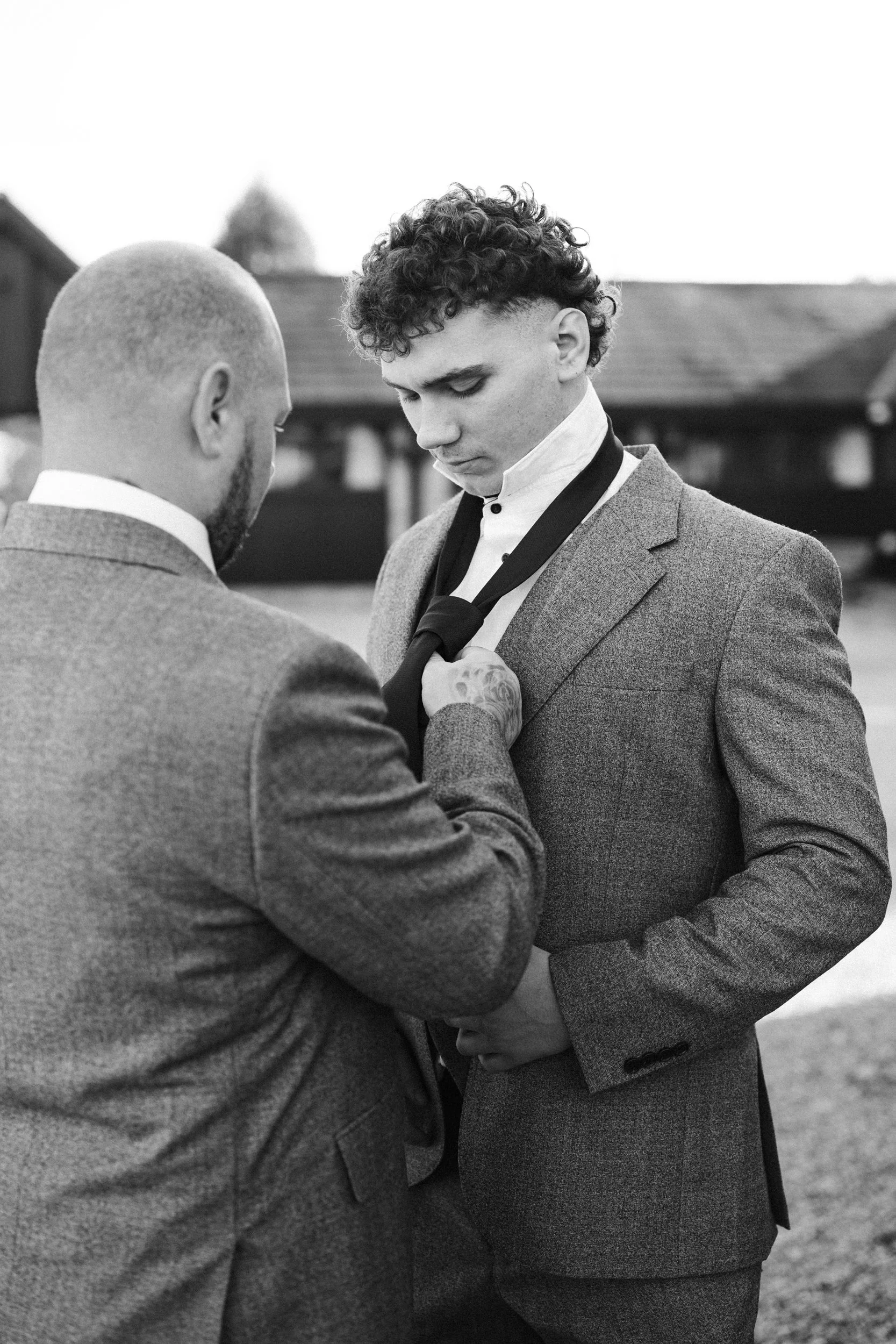 Two men in suits are outdoors, one helping the other adjust a tie. The man on the right has curly hair, and the man on the left has a shaved head. The background shows a building with a sloped roof.
