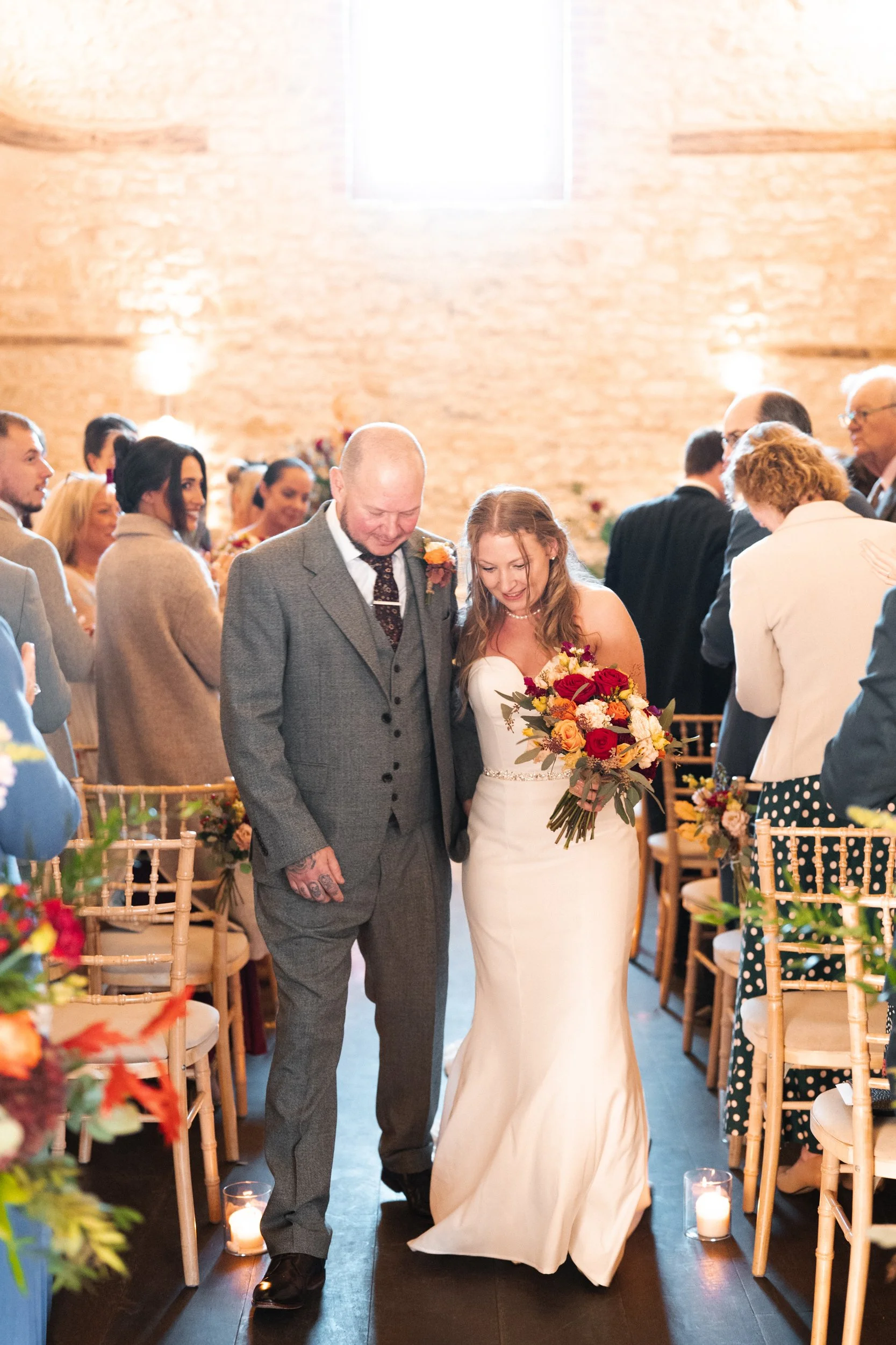 A bride in a white wedding gown holding a bouquet walking down the aisle with a man who appears to be her father. Guests are standing and clapping on either side in a warmly lit venue.
