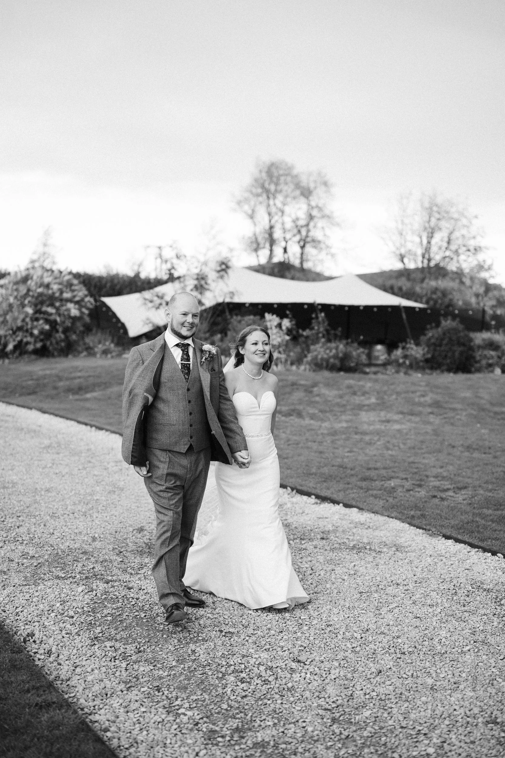 Black and white photo of a bride and groom walking hand-in-hand outdoors, with a tent and trees in the background.