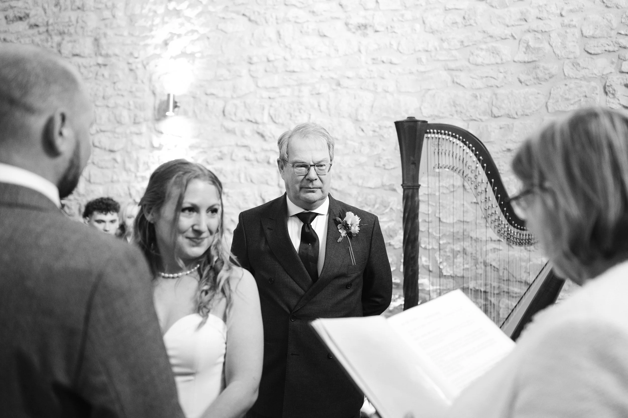 Black and white photo of a wedding ceremony with a bride, groom, officiant, and a man with a boutonniere, standing in front of a stone wall and a harp.
