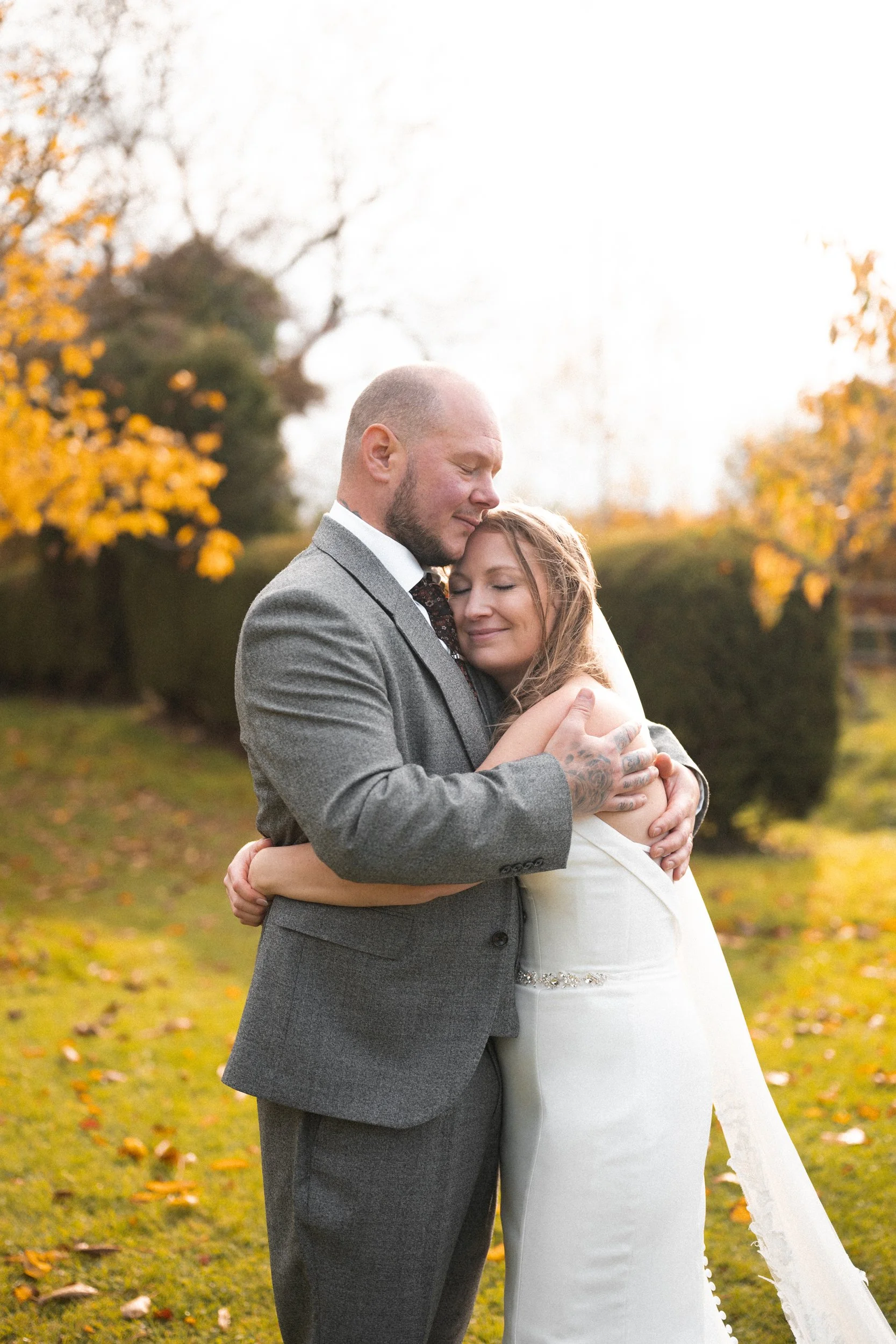 A couple embracing outdoors in autumn, with the man in a gray suit and the woman in a white gown, their eyes closed and happy, surrounded by trees with orange leaves.