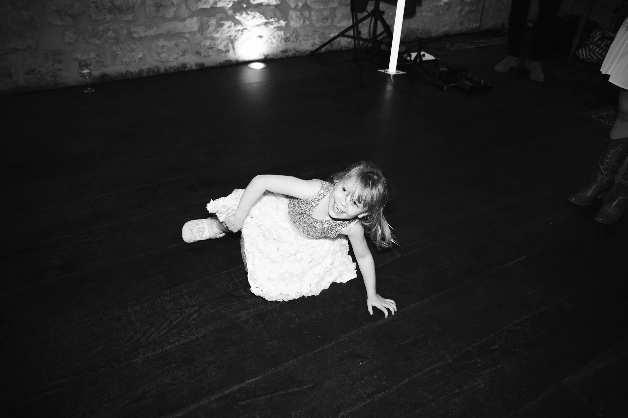 A young girl in a dress is on the floor, smiling and looking up, at what appears to be an indoor event with a stone wall background.