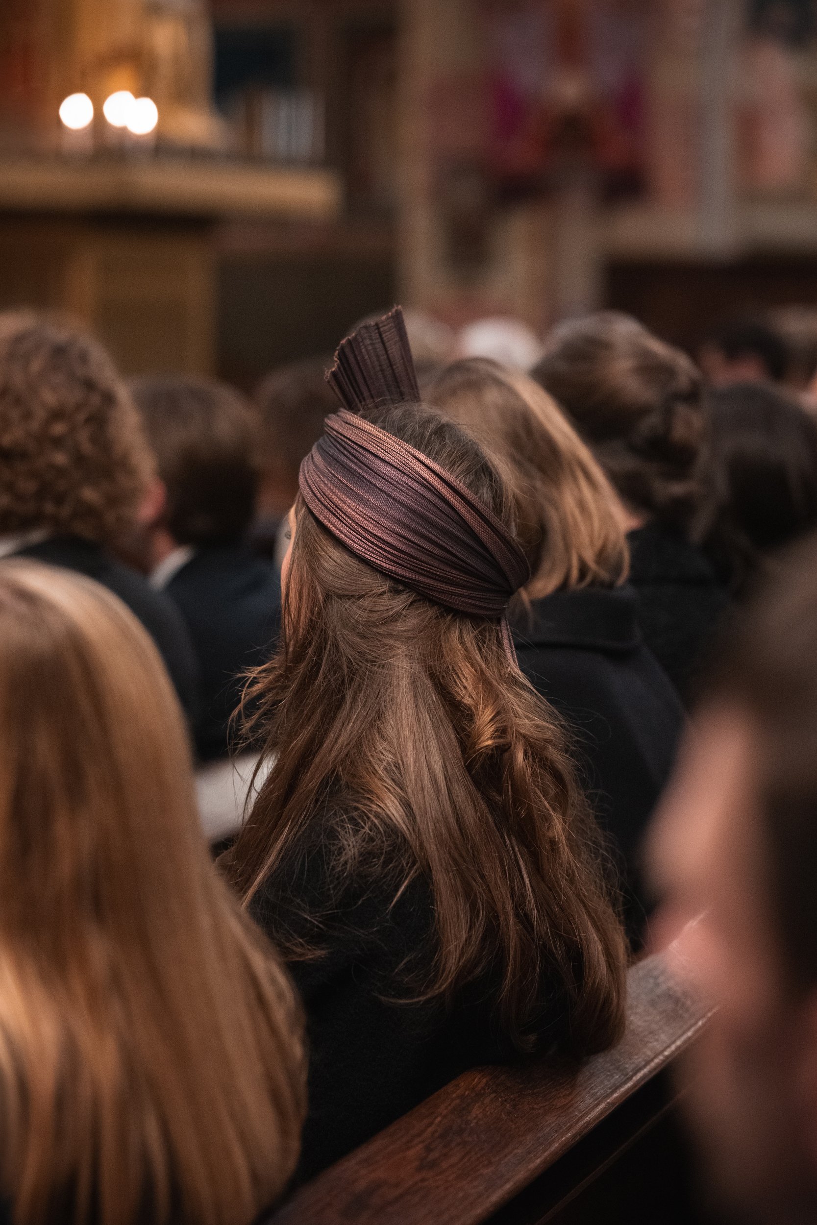 A woman with long, wavy brown hair wearing a brown head wrap, sitting in a crowded indoor setting, possibly a church or lecture hall, with other people around her.