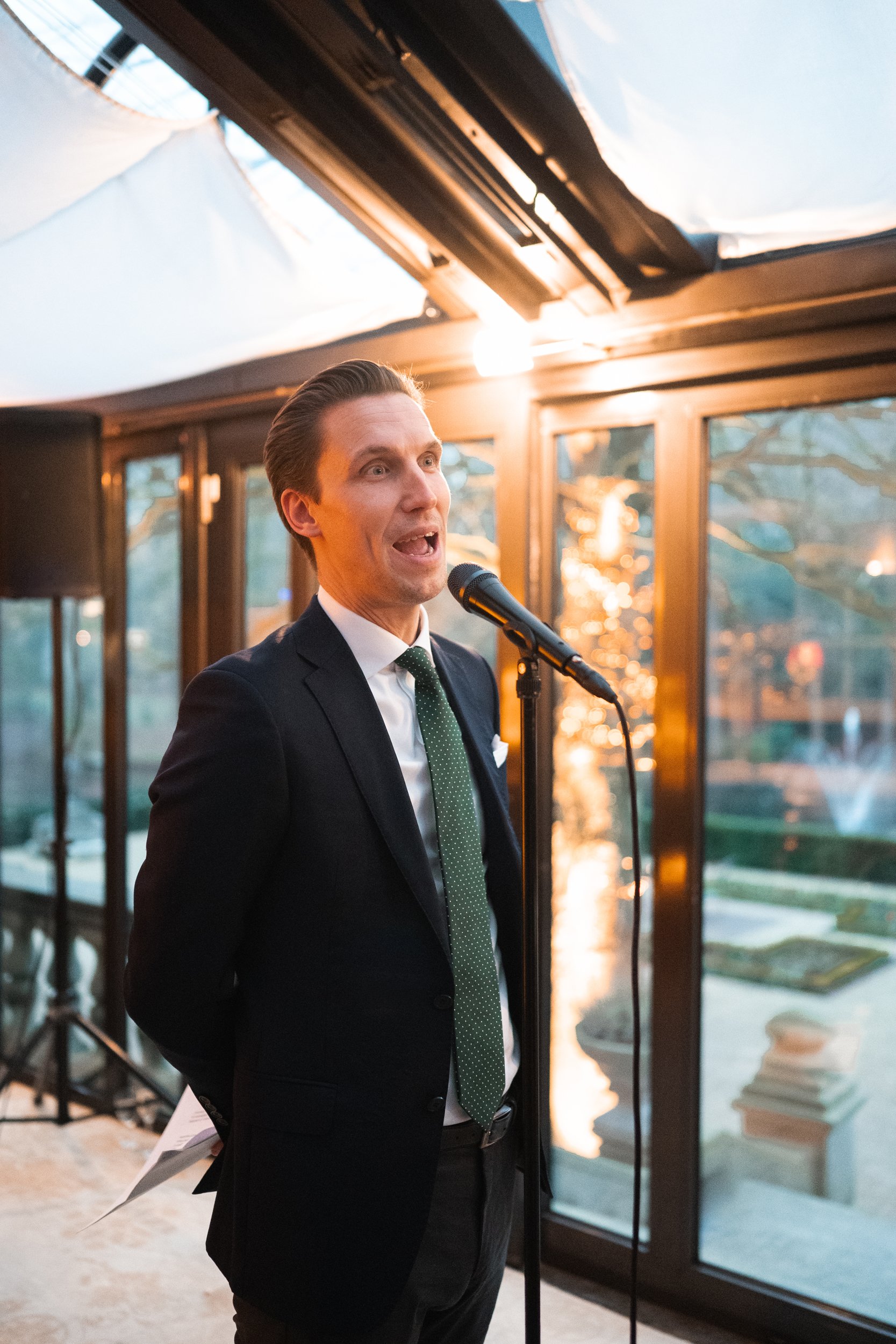 A man in a suit speaking into a microphone during an indoor event with large windows and outdoor scenery visible.