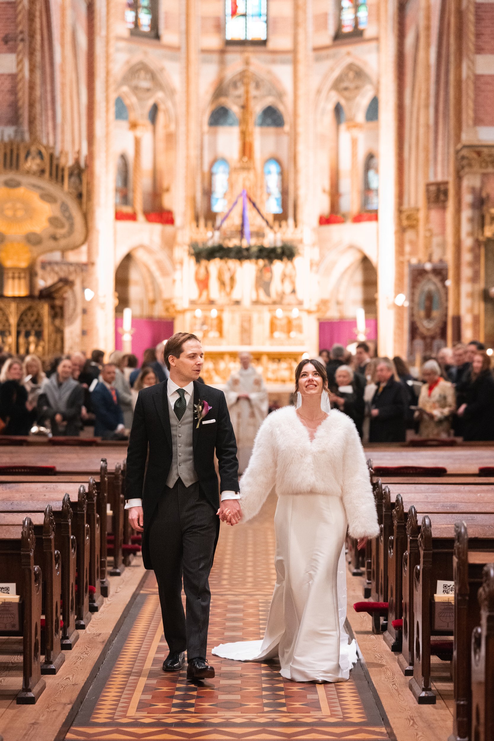 A bride and groom holding hands and walking down the aisle inside a church, with the bride looking up and smiling. The background shows guests seated and a decorated altar with religious icons and stained glass windows.