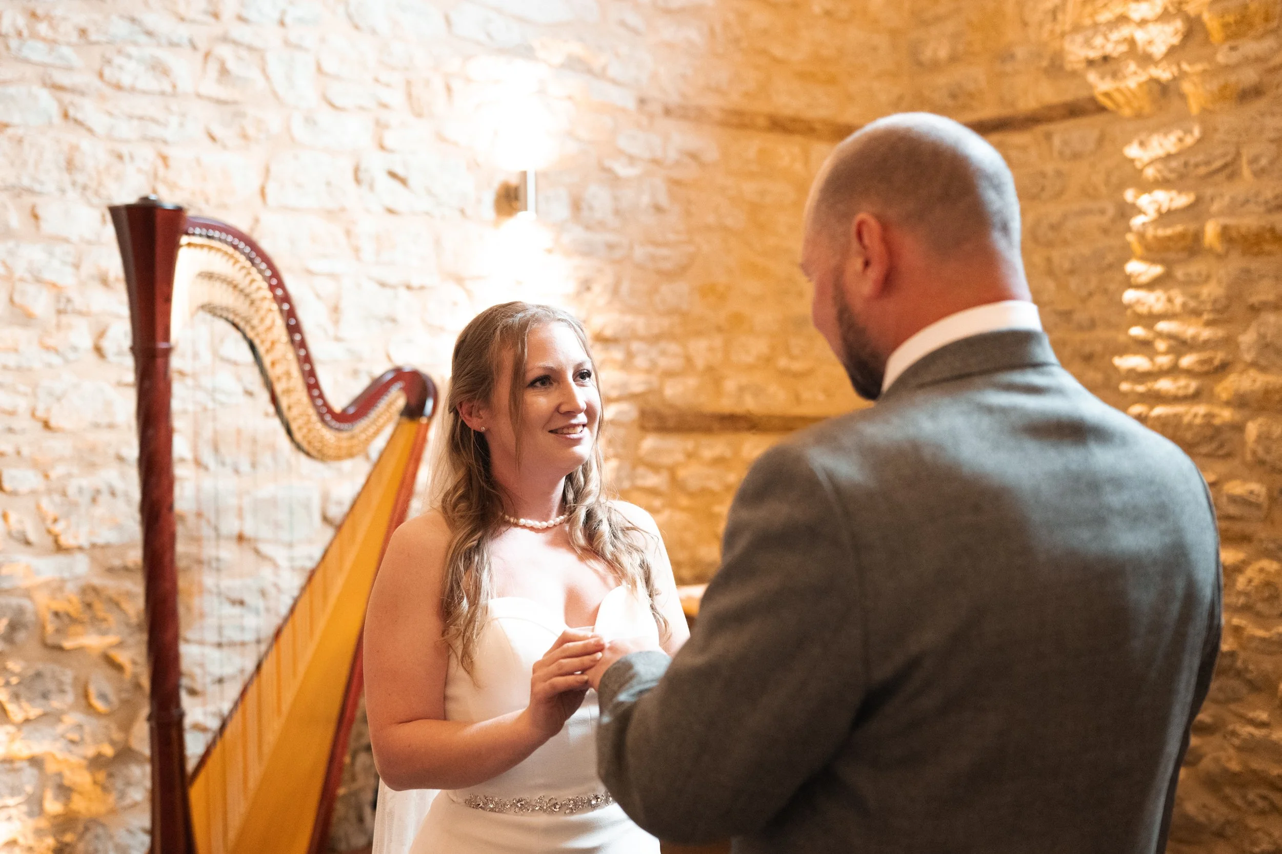 A bride and groom during their wedding ceremony, holding hands and gazing at each other inside a rustic brick-walled room with a harp in the background.