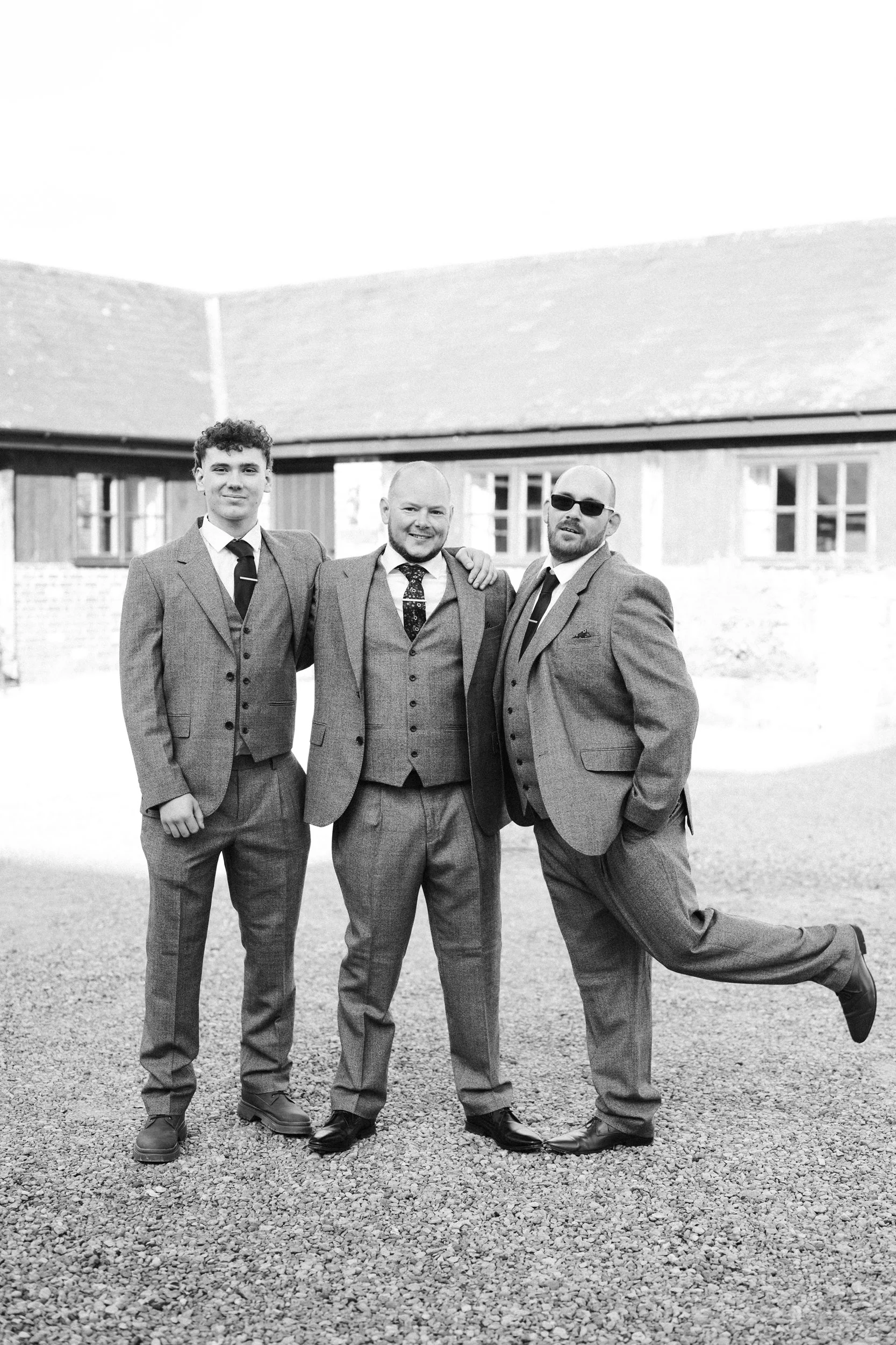 Three men in suits posing together outdoors in front of a building, smiling, with one man in sunglasses lifting one leg.