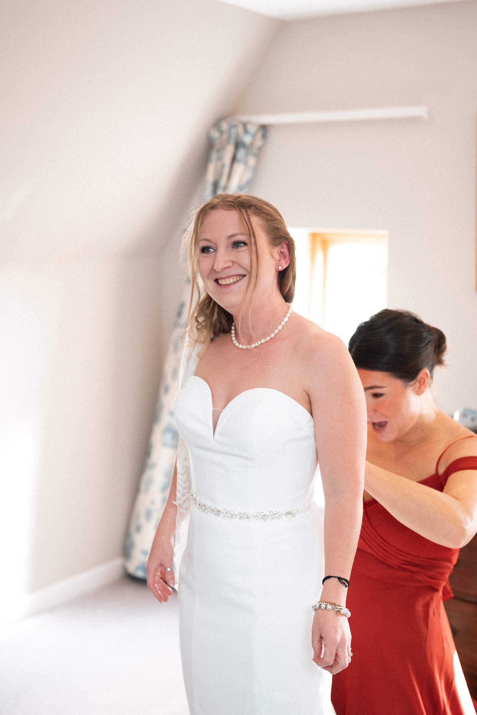A bride in a strapless white wedding dress with a pearl necklace, smiling as she is helped with her dress by a woman in a red dress.
